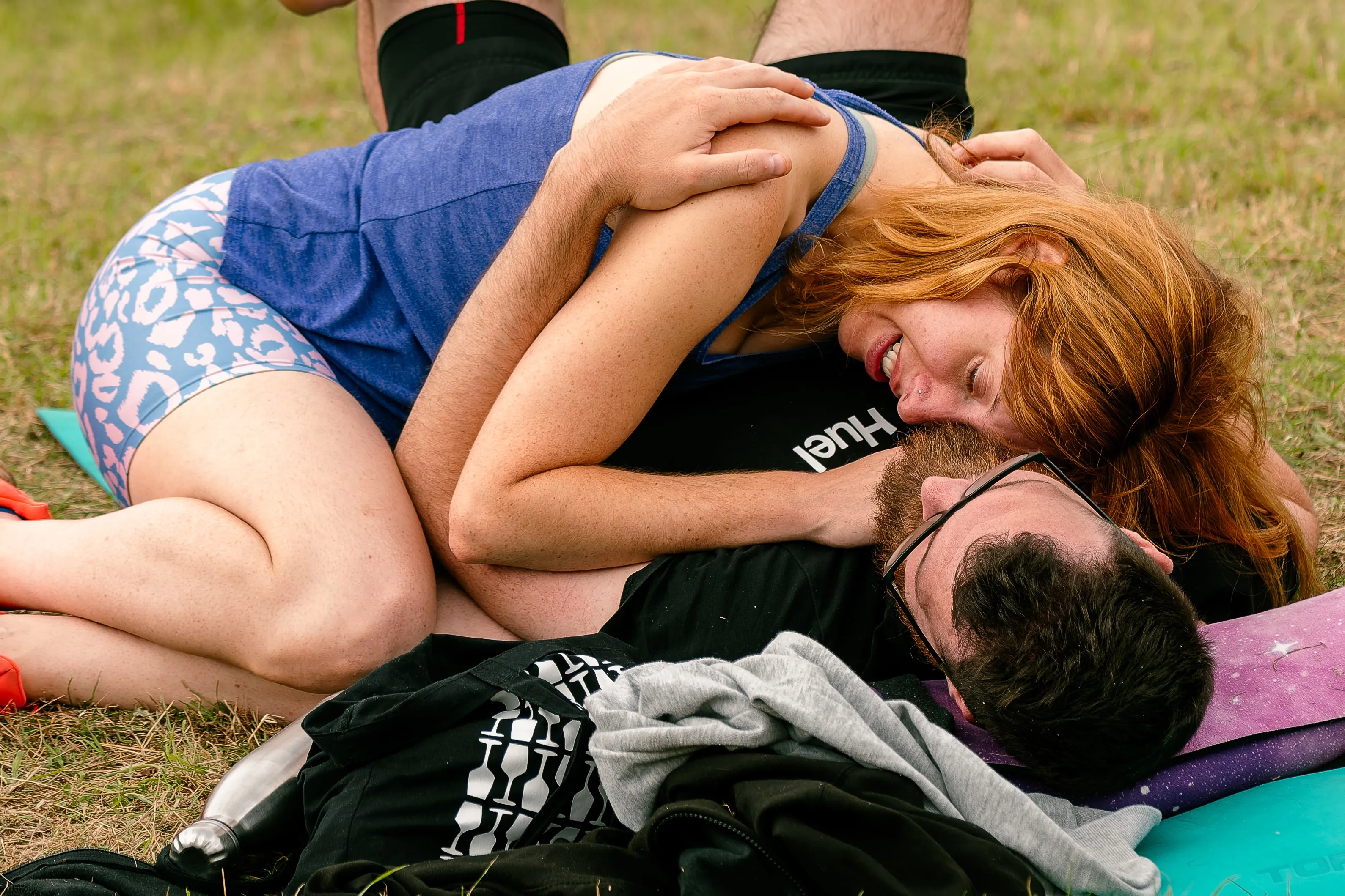 A couple lying on the grass, a red-haired woman leaning over a bearded man as they laugh together.