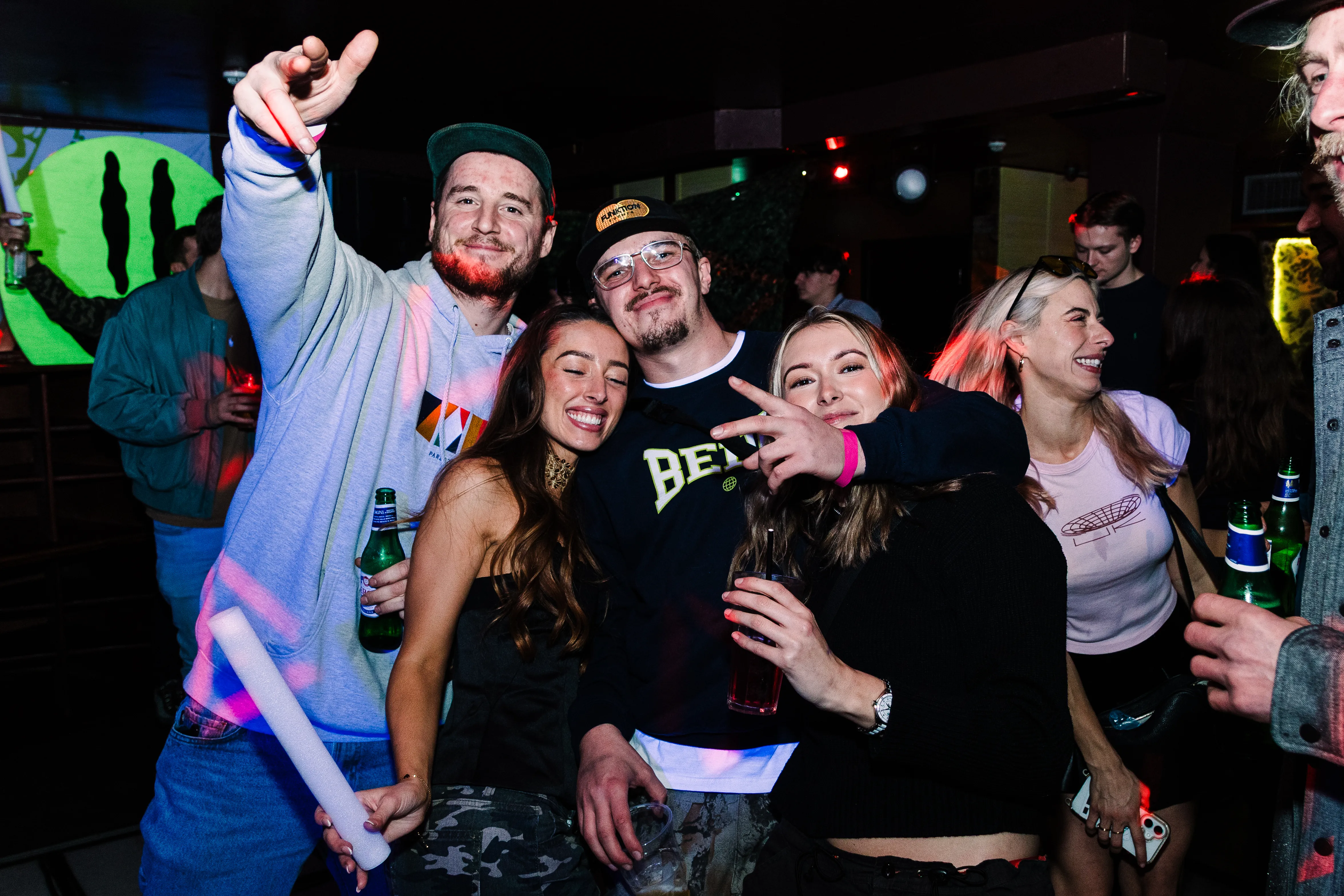 A group of friends posing together with a glowstick raised, lit by camera flash in a dark club.