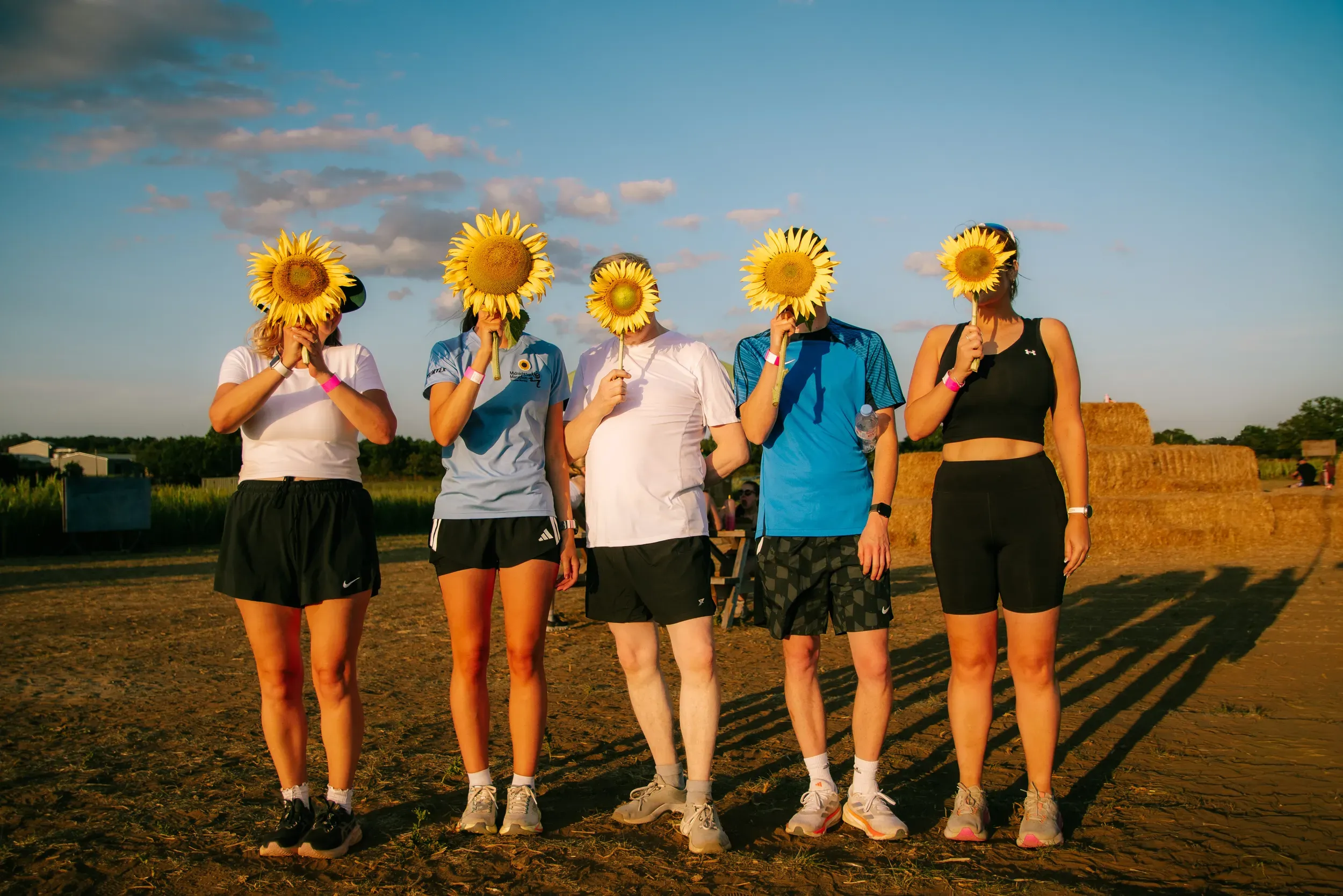 Five runners lined up holding sunflowers over their faces, long shadows across the ground in the last of the sun.