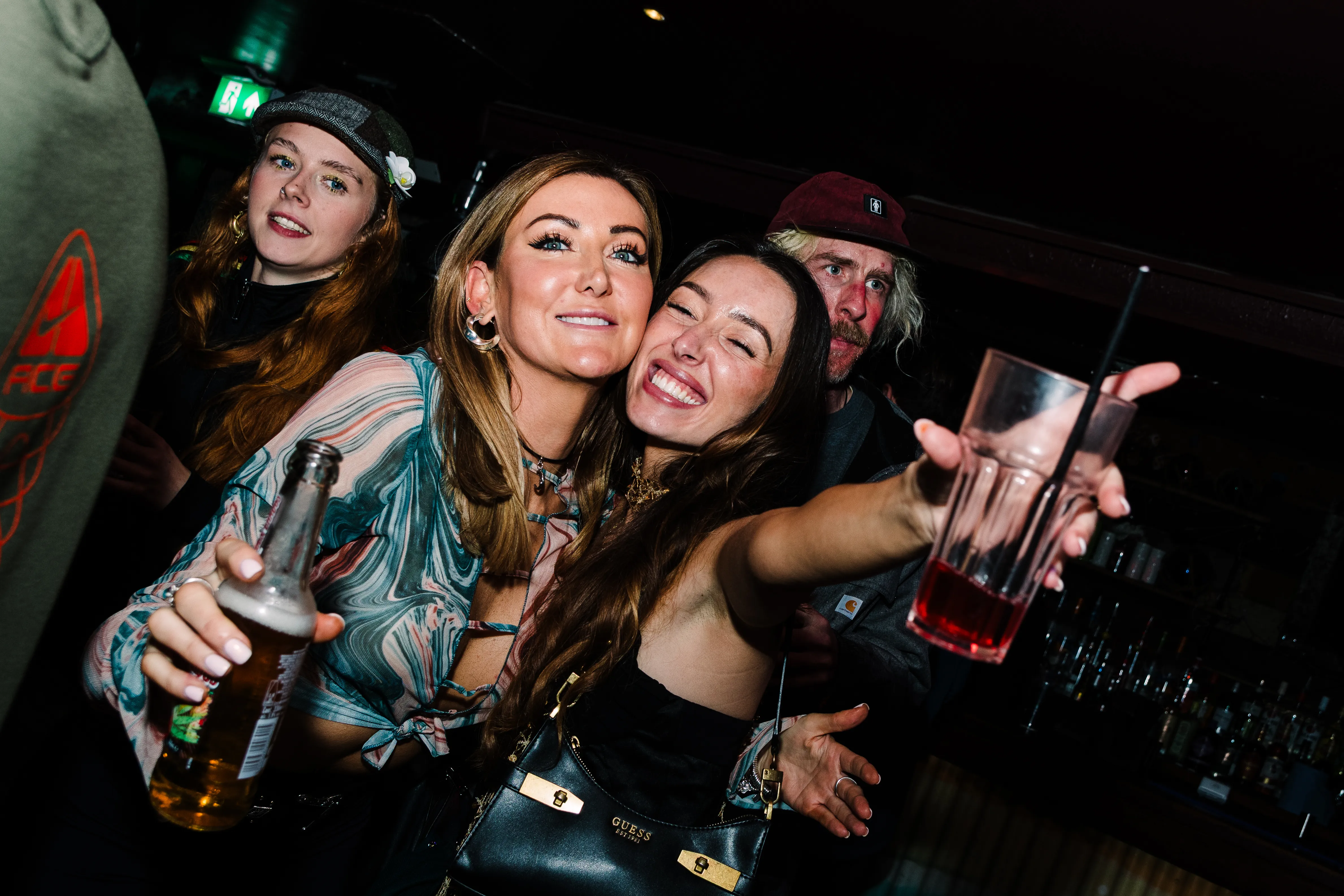 Three friends laughing at the bar, one reaching towards the camera past a tall drink.