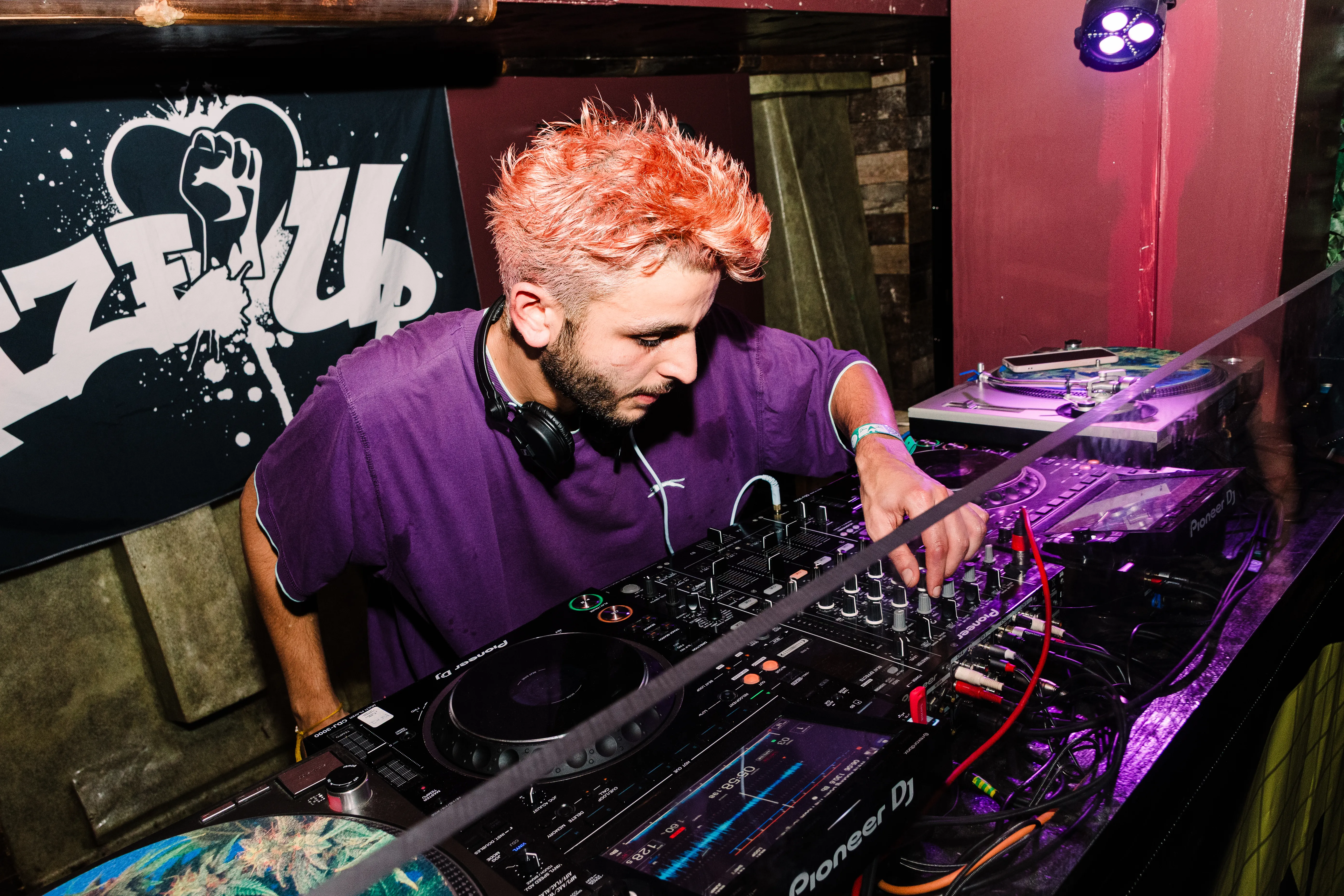 A DJ with bright orange hair in a purple shirt working the decks under club lighting.