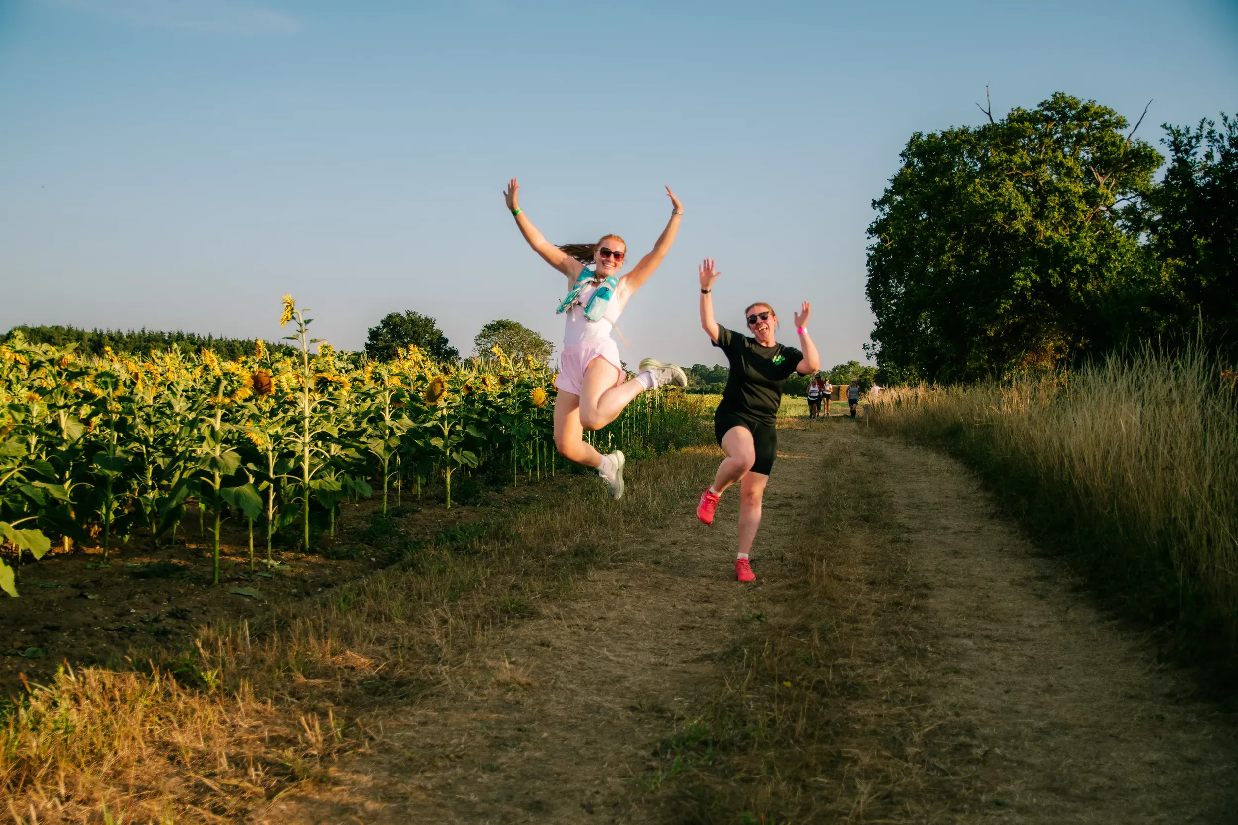 Two runners jumping in the air side by side on the track, sunflowers to their left and golden light behind.