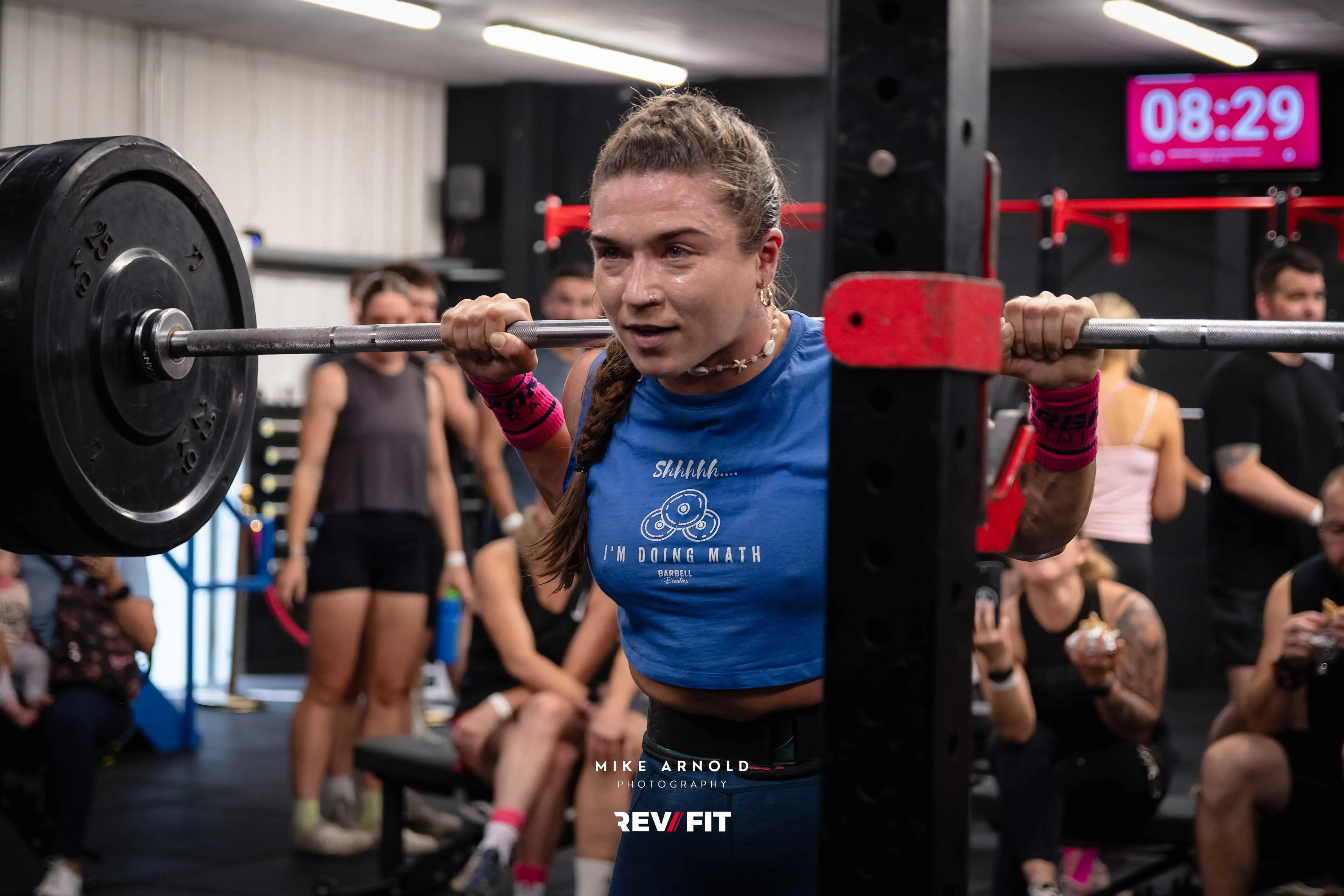 A competitor in a blue team shirt squatting a loaded barbell as the crowd cheers behind.
