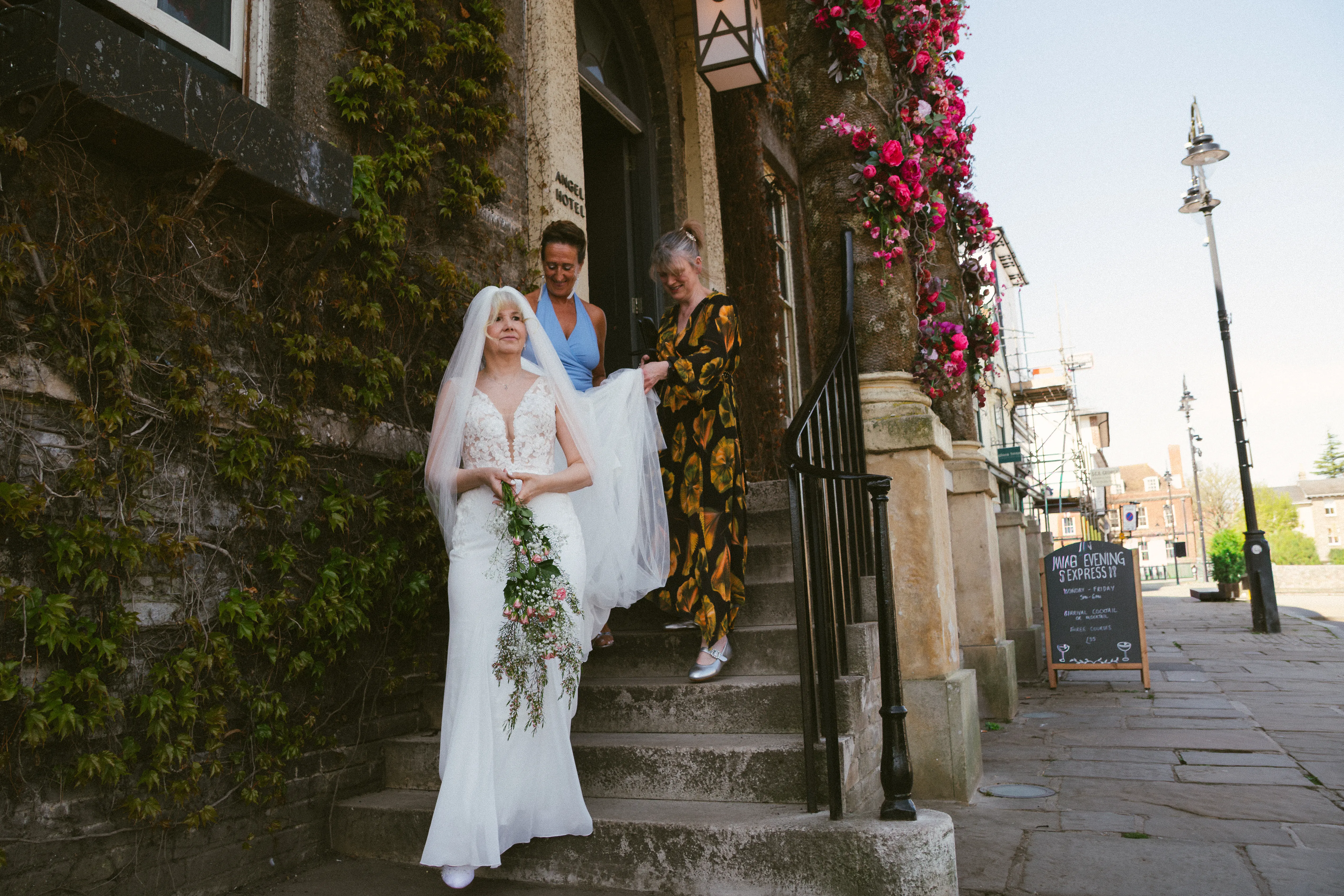 A bride in a lace gown holding a bouquet, walking down stone steps outside an ivy-covered building.