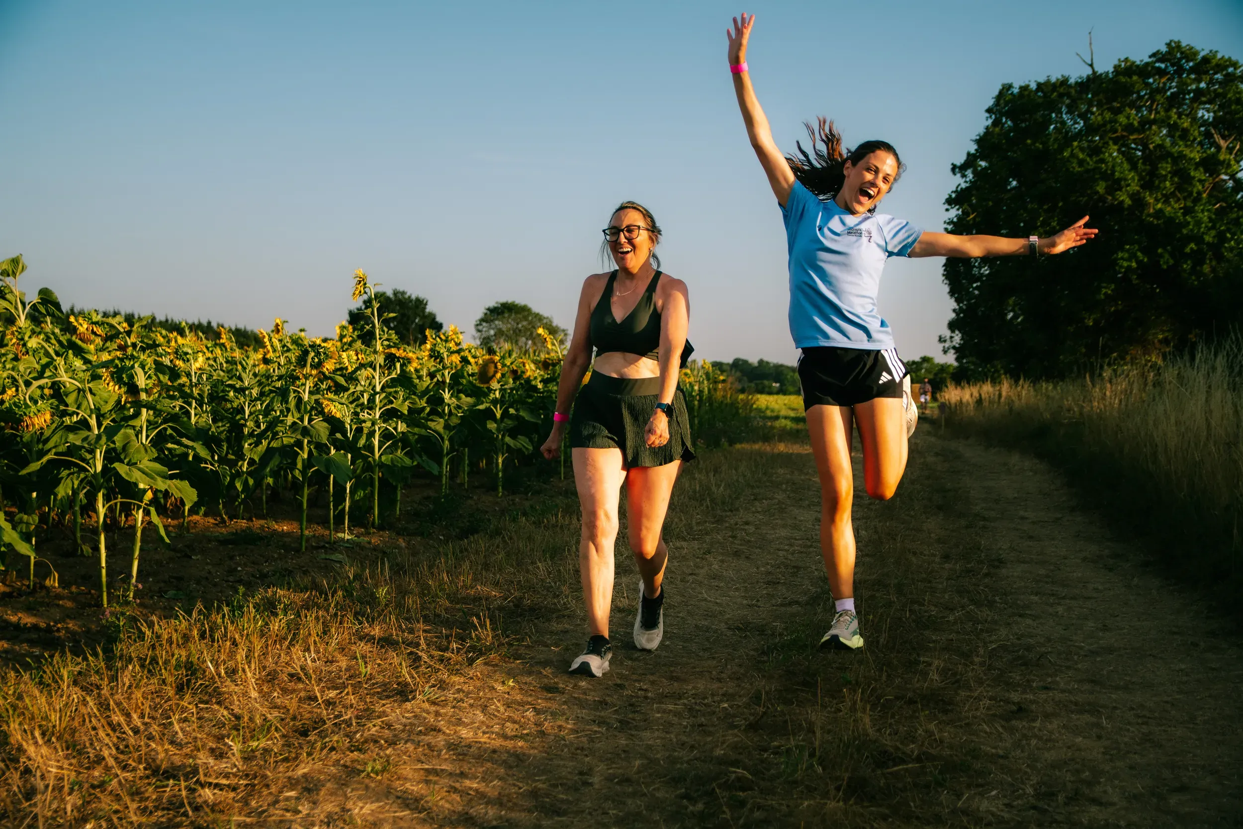 A runner leaping into the air with her arms out beside the sunflower field, her friend laughing beside her.