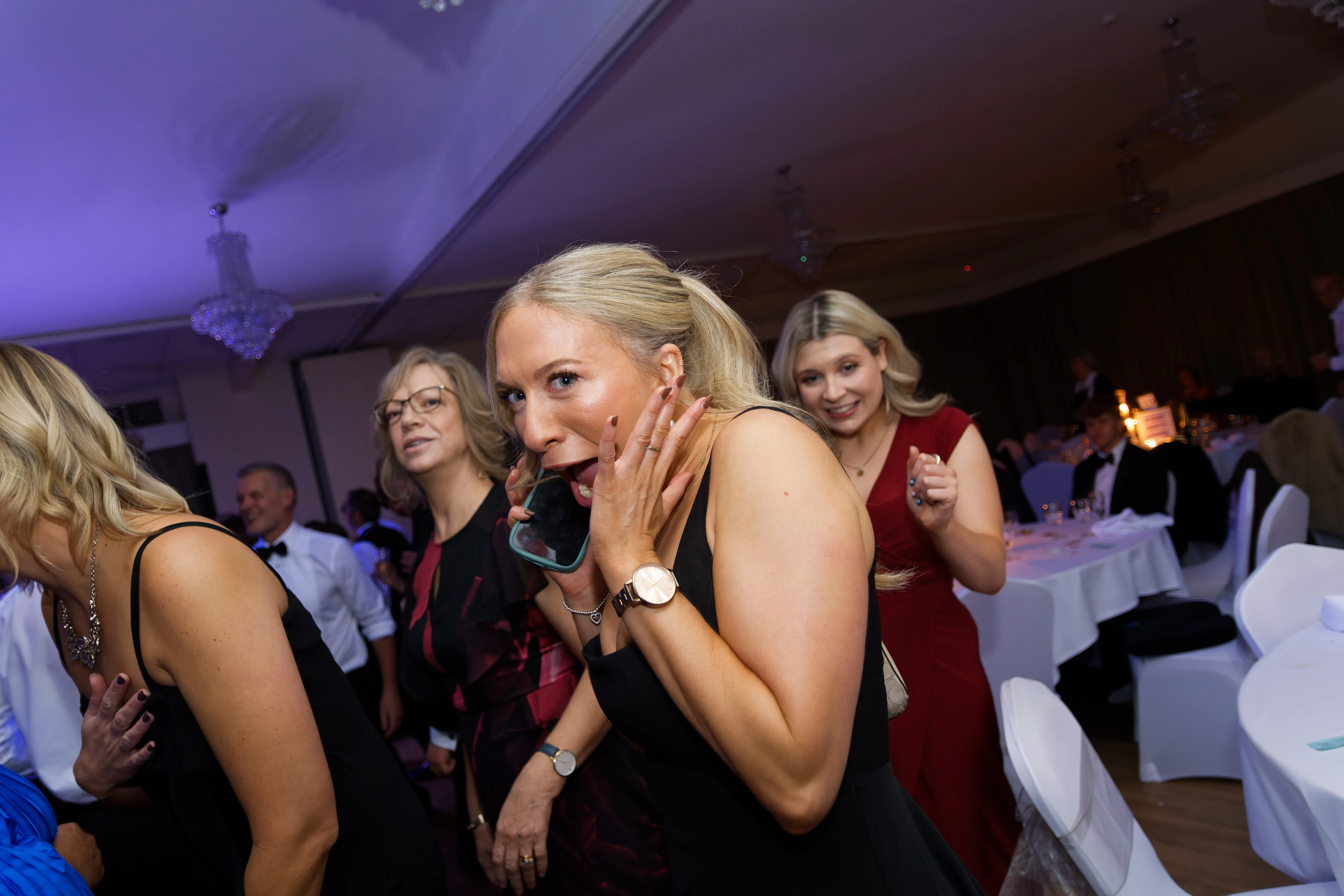 A woman in a black dress reacting with mock surprise, hand to her face.