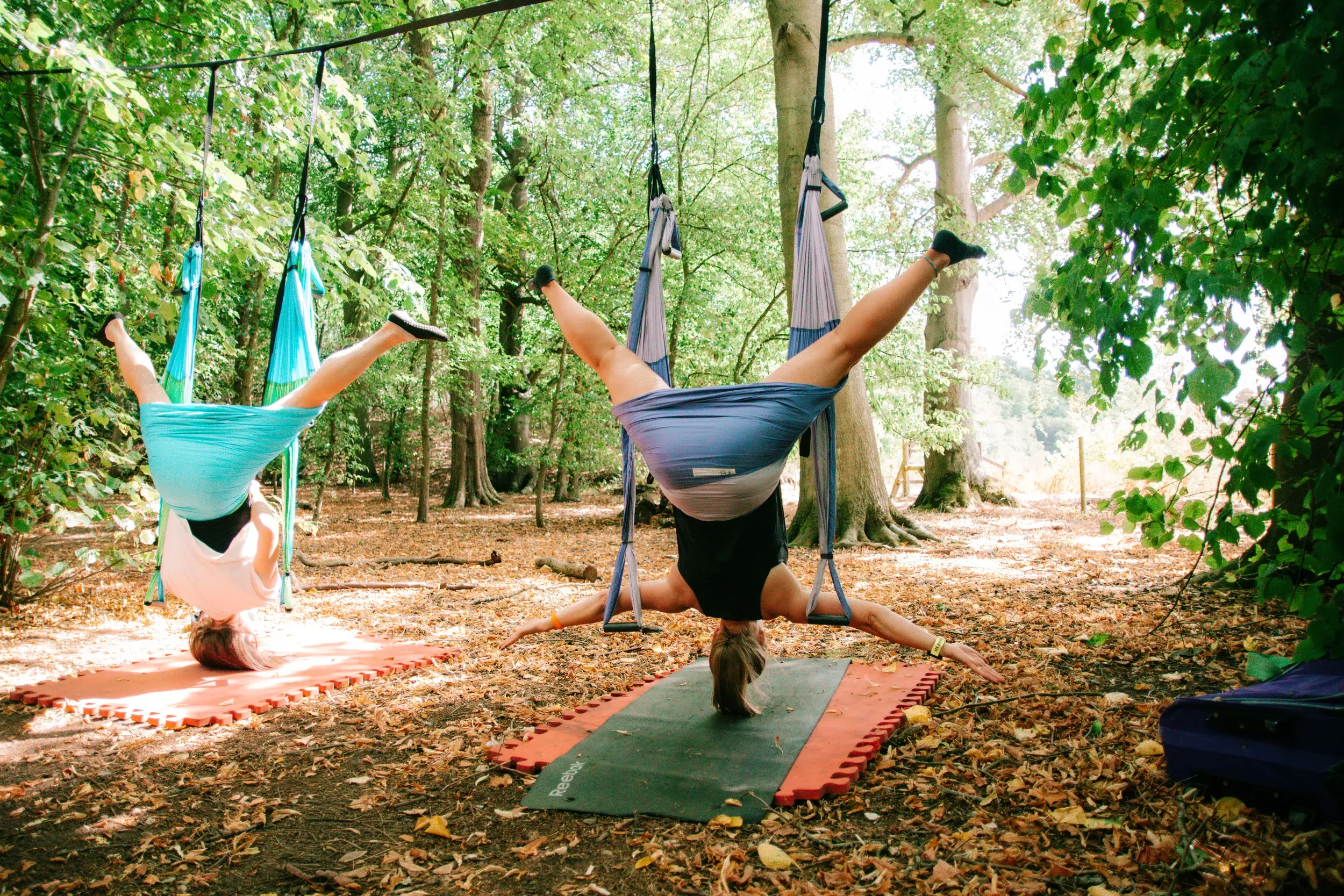 Two people hanging upside down in aerial yoga hammocks among the trees.