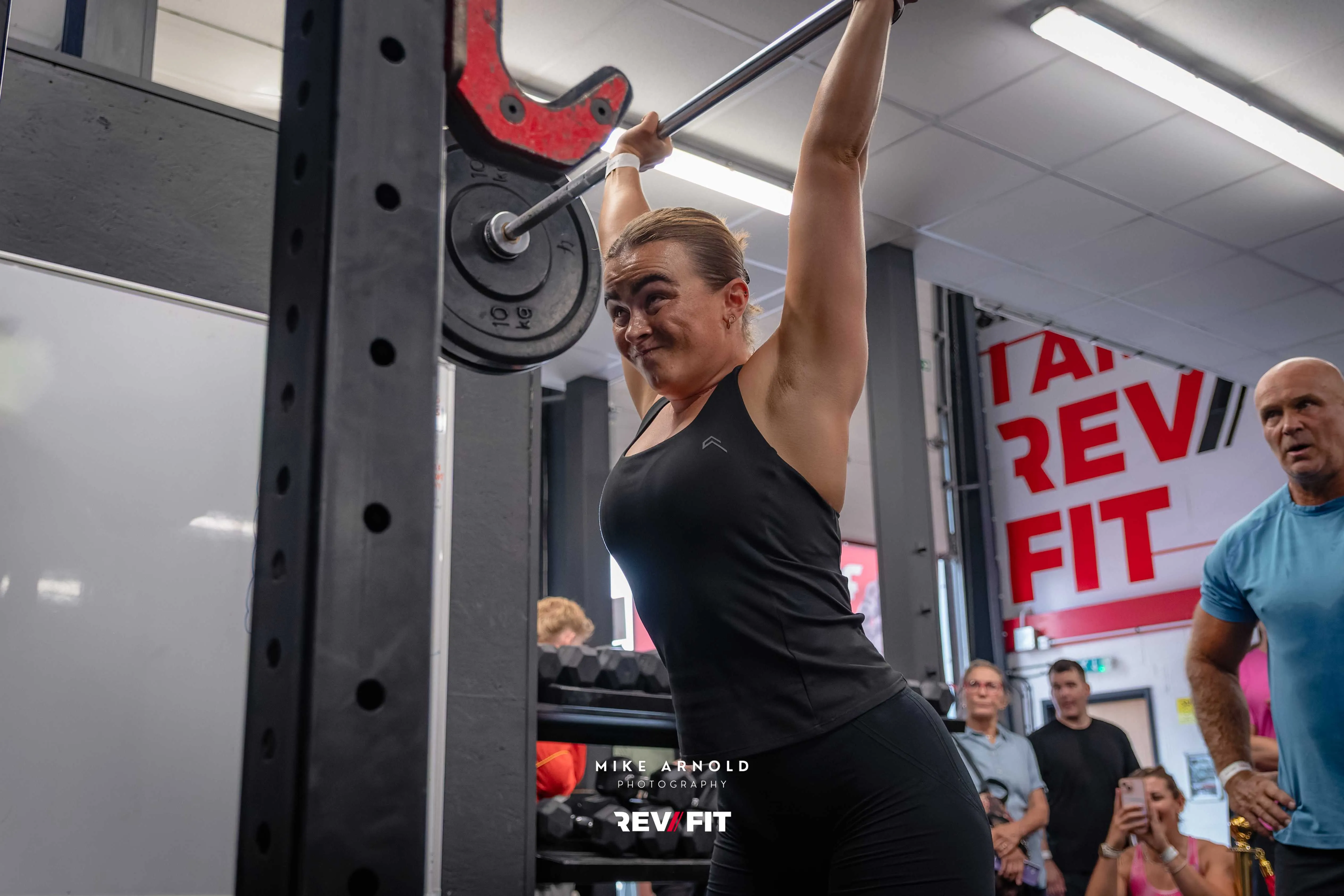 A competitor pressing a barbell overhead on the rig, supporters watching from the side.