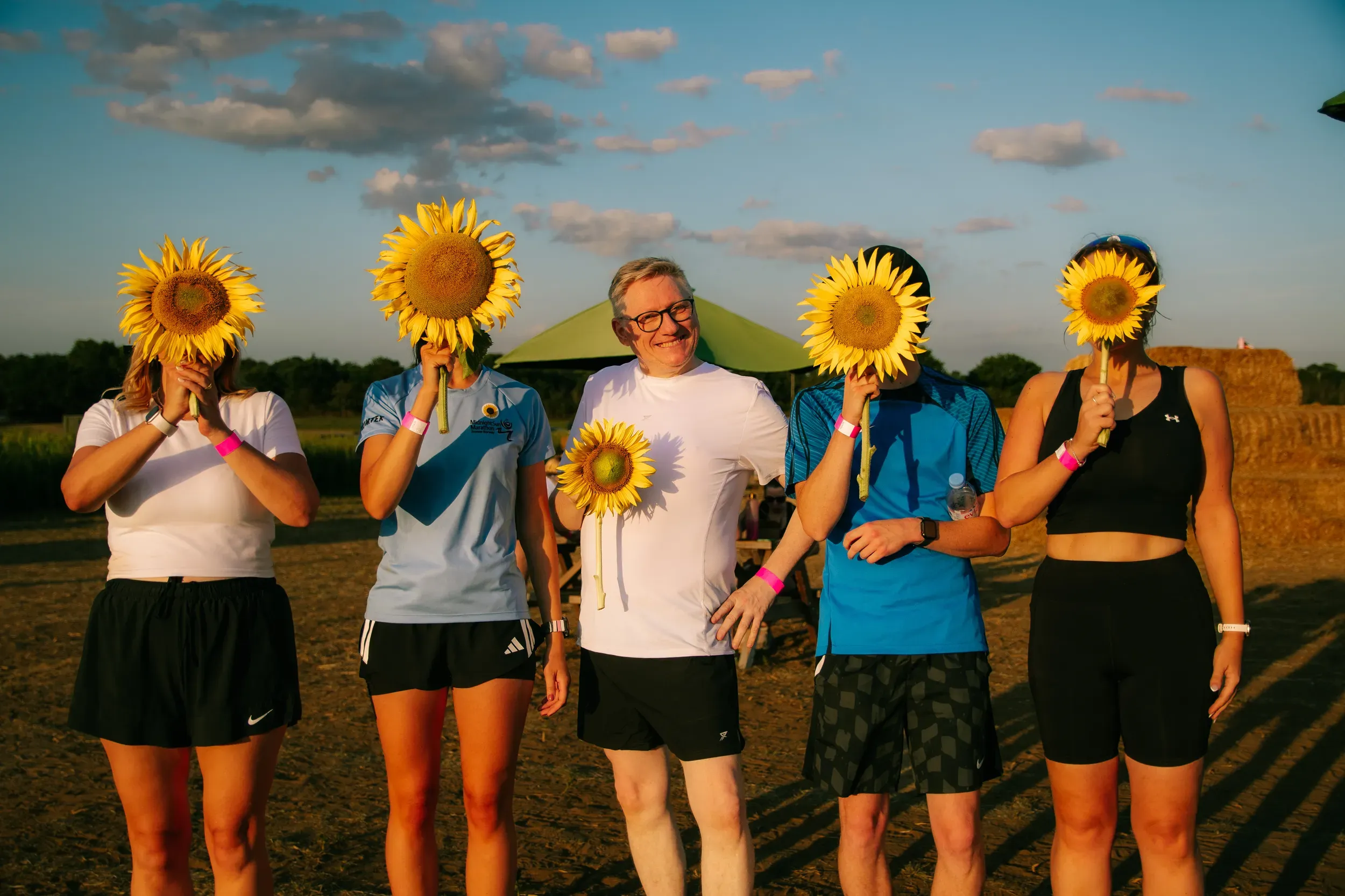 Five runners holding sunflowers up in front of their faces after the run, hay bales behind them.