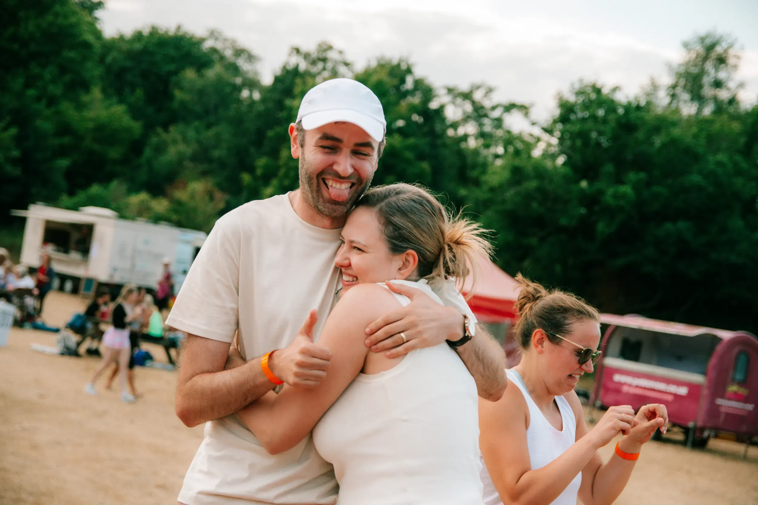 A man in a white cap pulling a friend into a hug by the food trucks, both of them laughing.