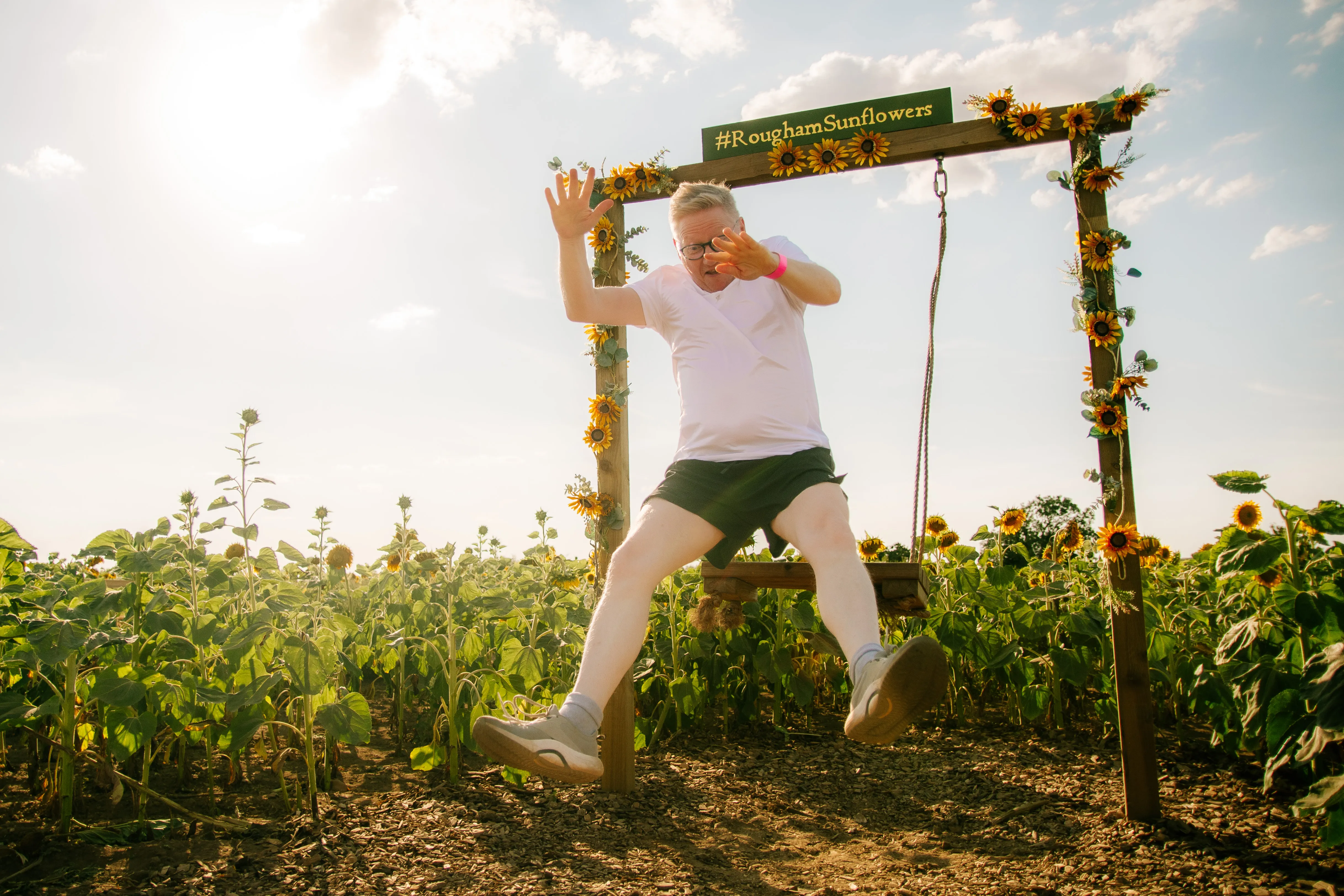A runner larking about on the swing under the #RoughamSunflowers sign, mid-air with the sun behind him.