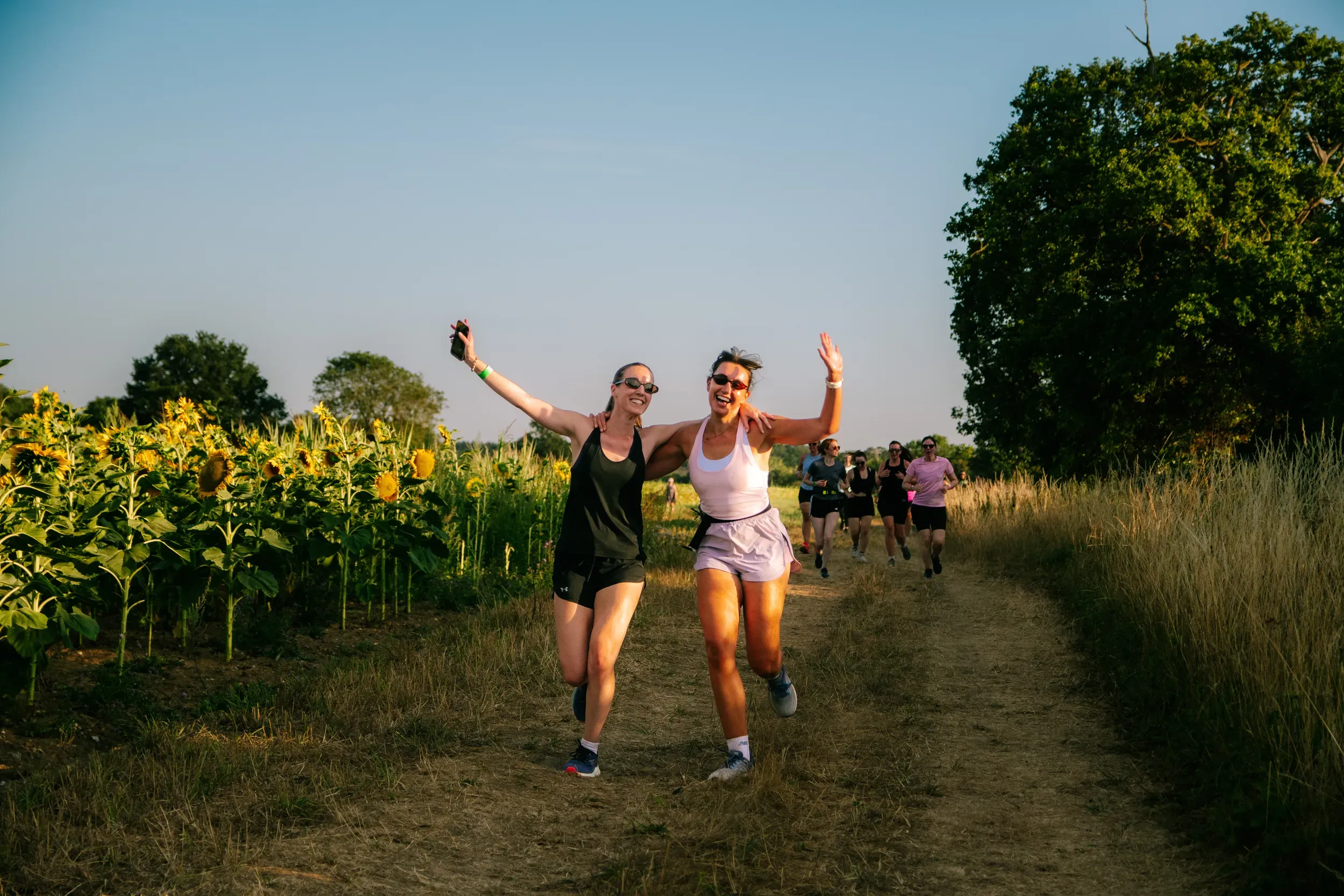 Two runners with their arms flung out, laughing, the rest of the group behind them along the sunflower row.