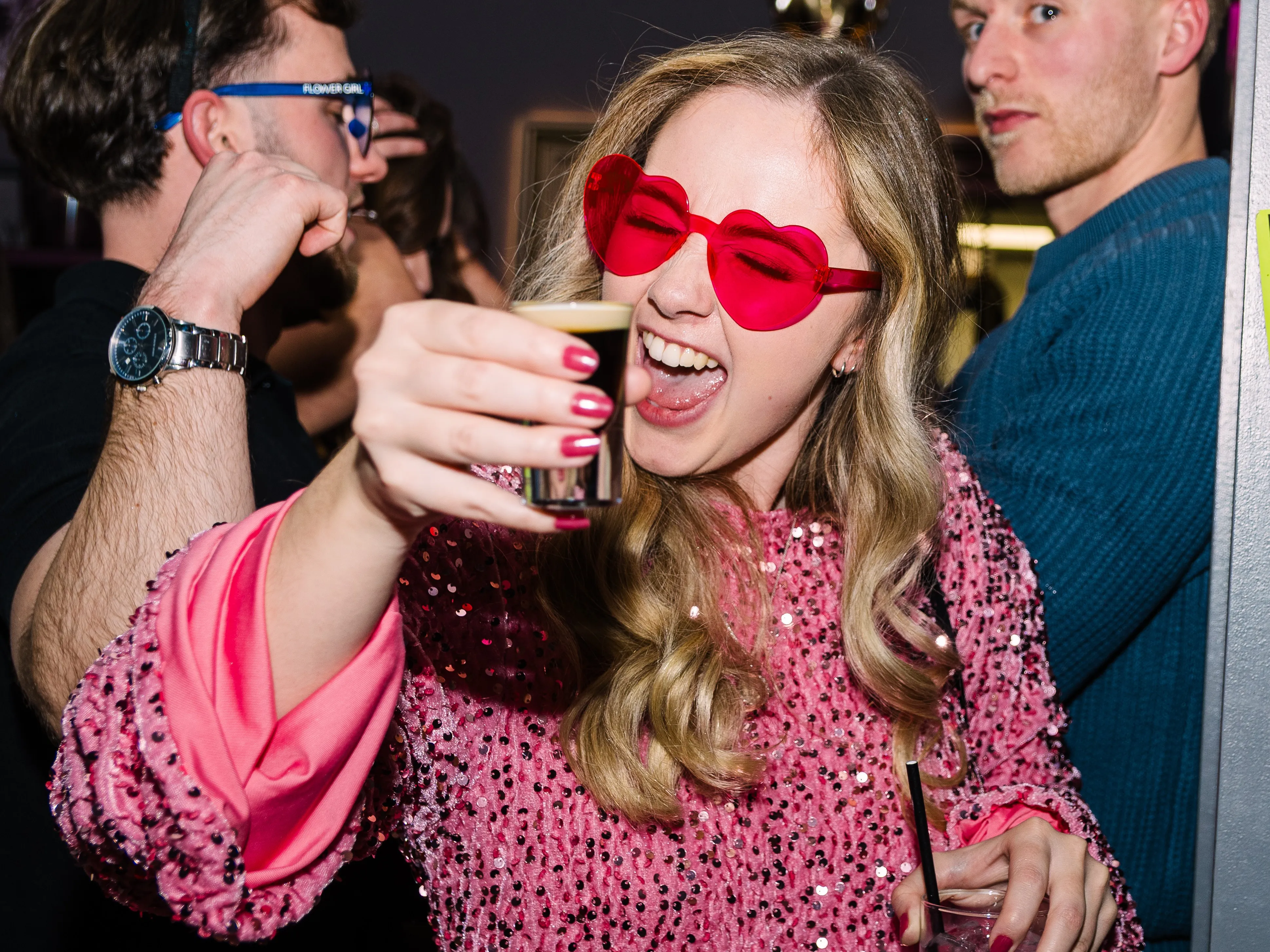 A woman in a pink sequined top and heart-shaped sunglasses laughing as she holds up a shot glass.