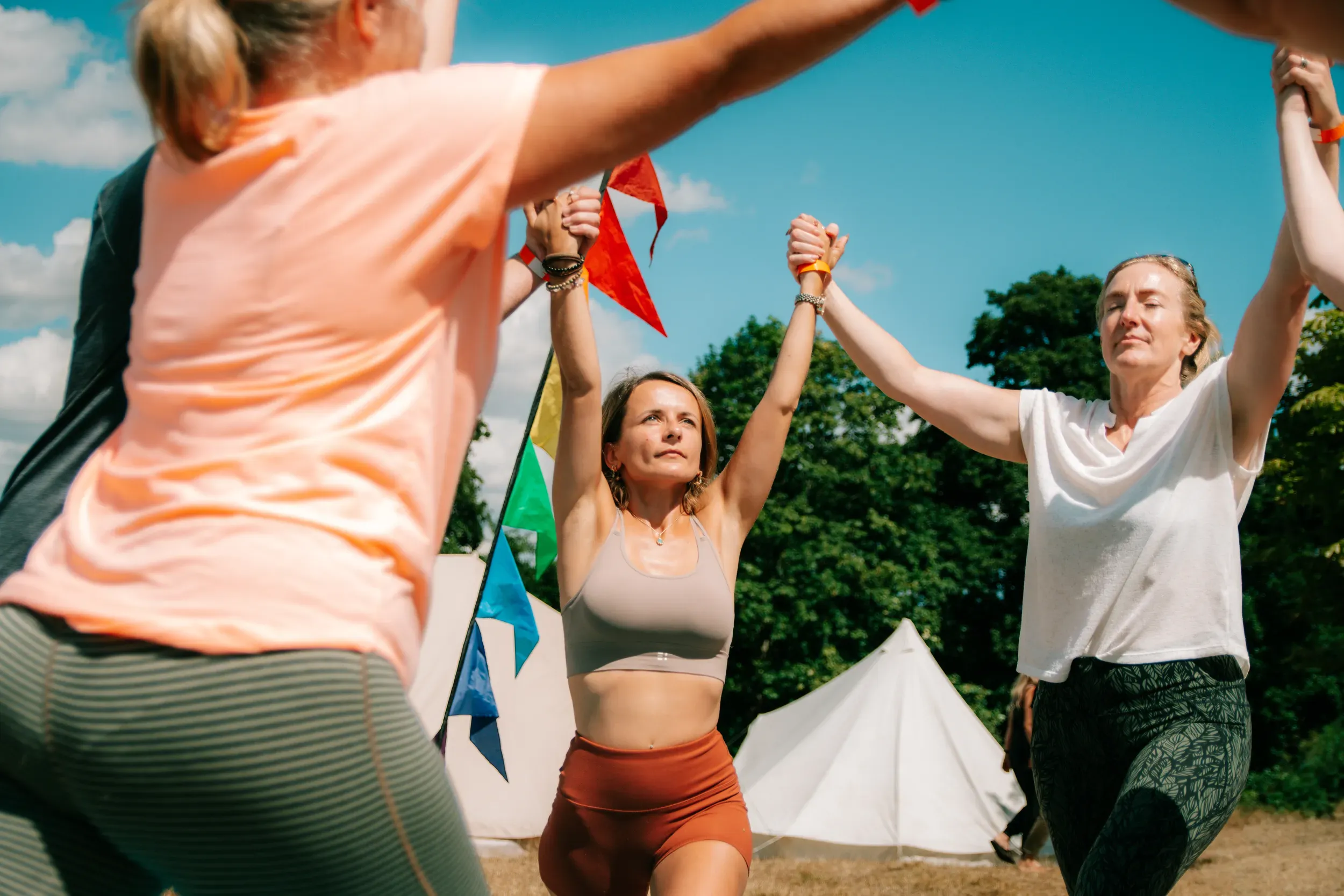 A group holding hands aloft in a circle under a bright blue sky, festival bunting and bell tents behind them.