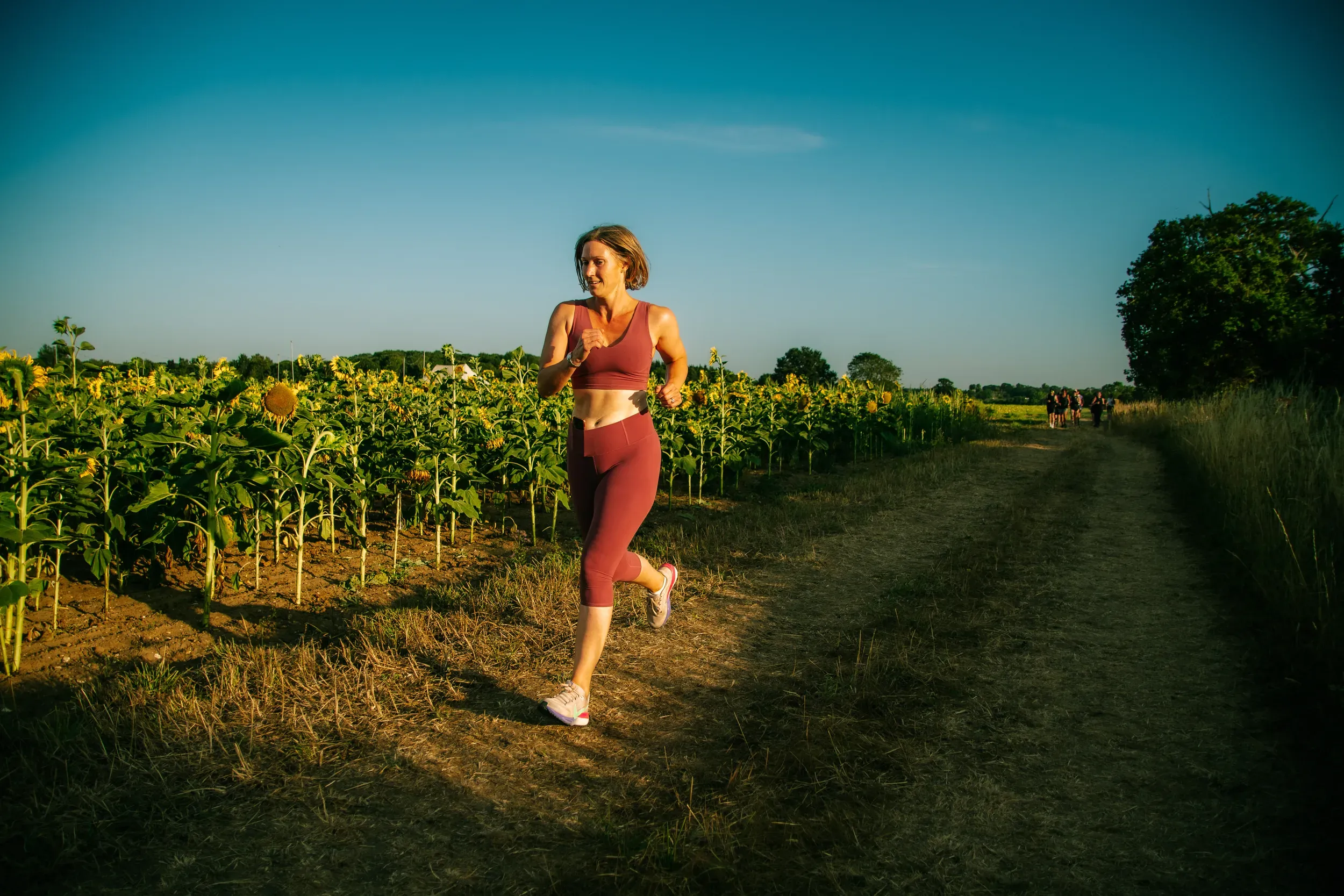 A runner in dusty pink alongside a full row of sunflowers, the track stretching away behind her.