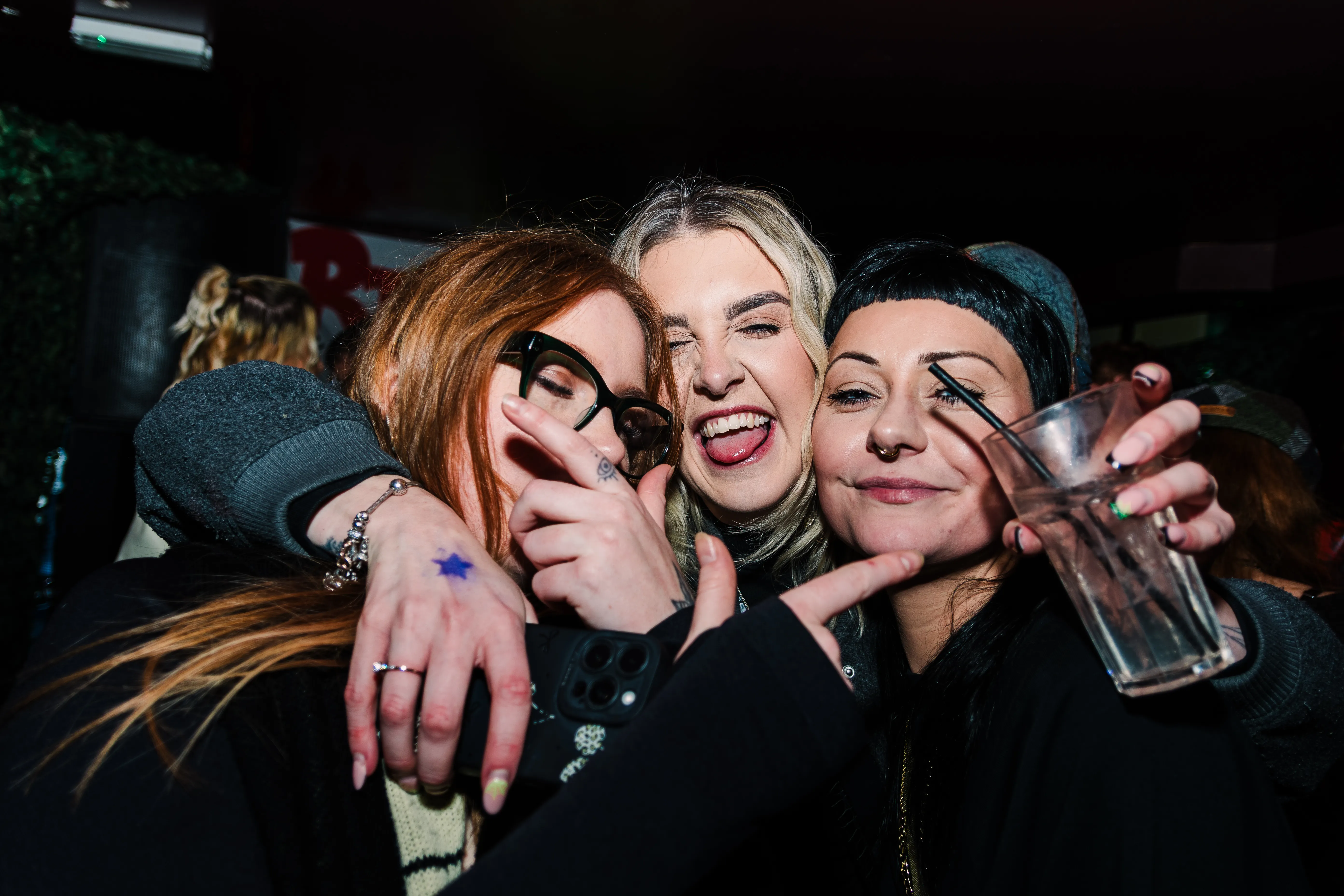 Three friends squeezed into a hug, tongues out, drinks in hand.