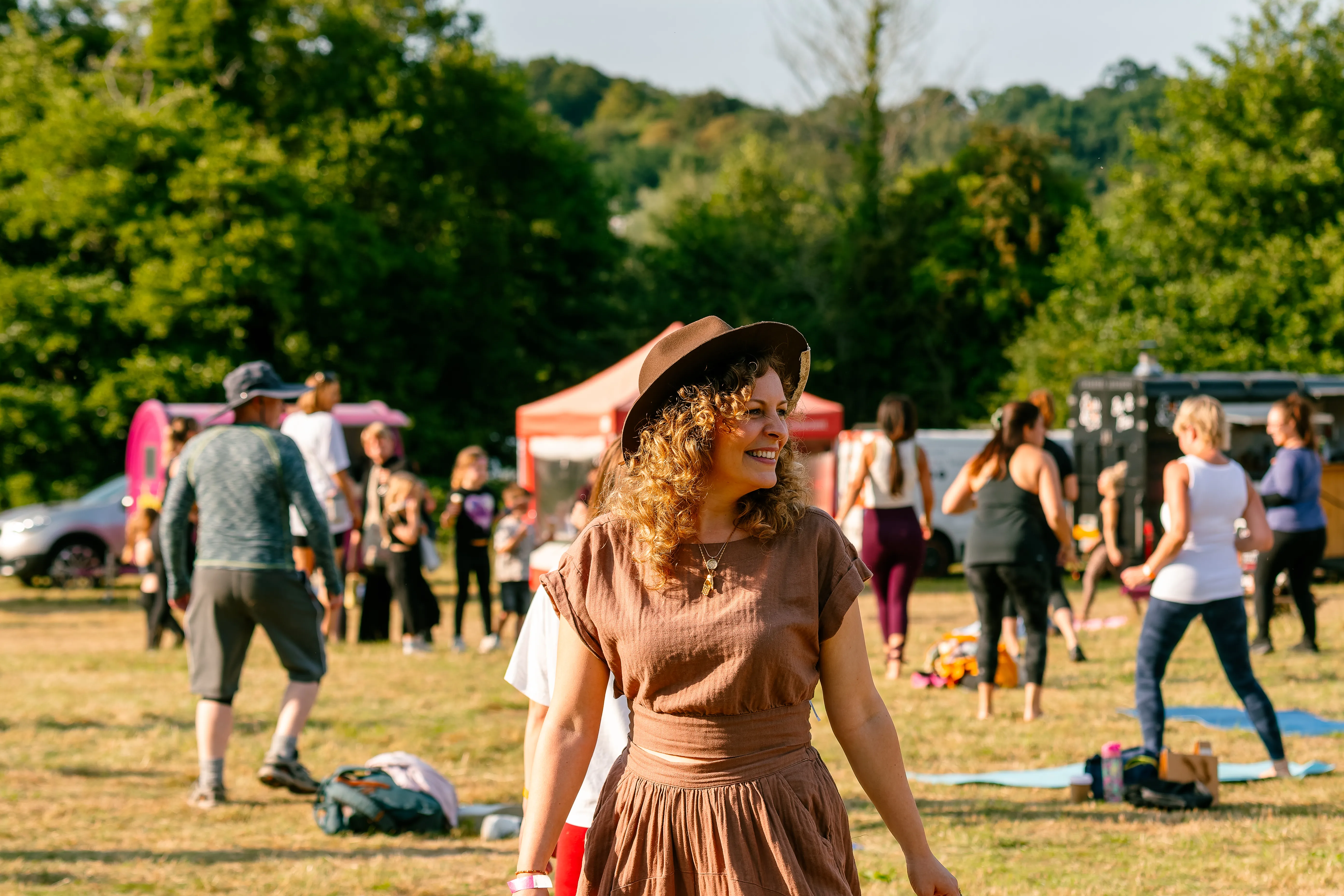 A woman in a brown wide-brimmed hat and earth-toned dress smiling in the evening sun, festival stalls behind her.