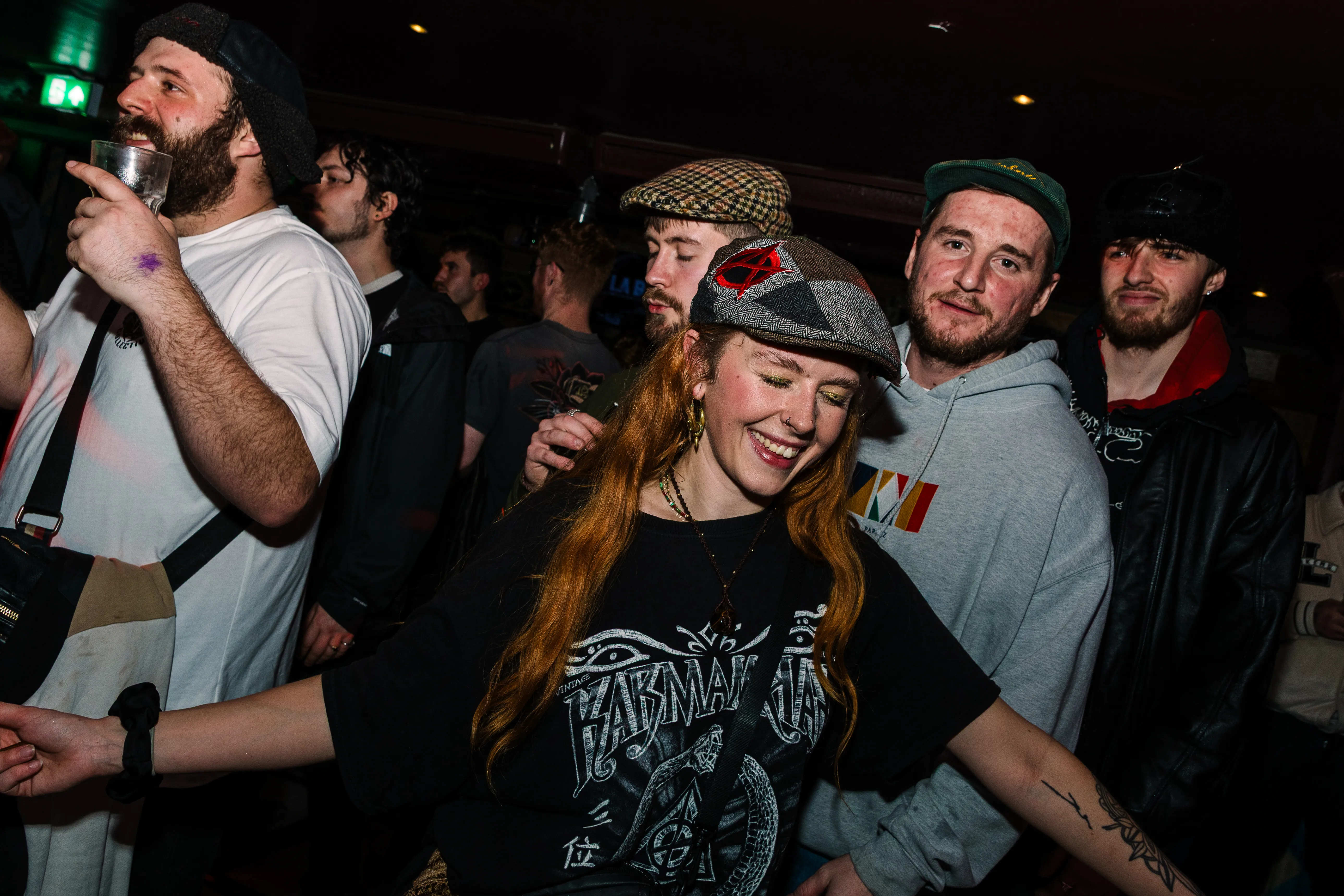 A smiling red-haired woman among friends in caps at a night event.