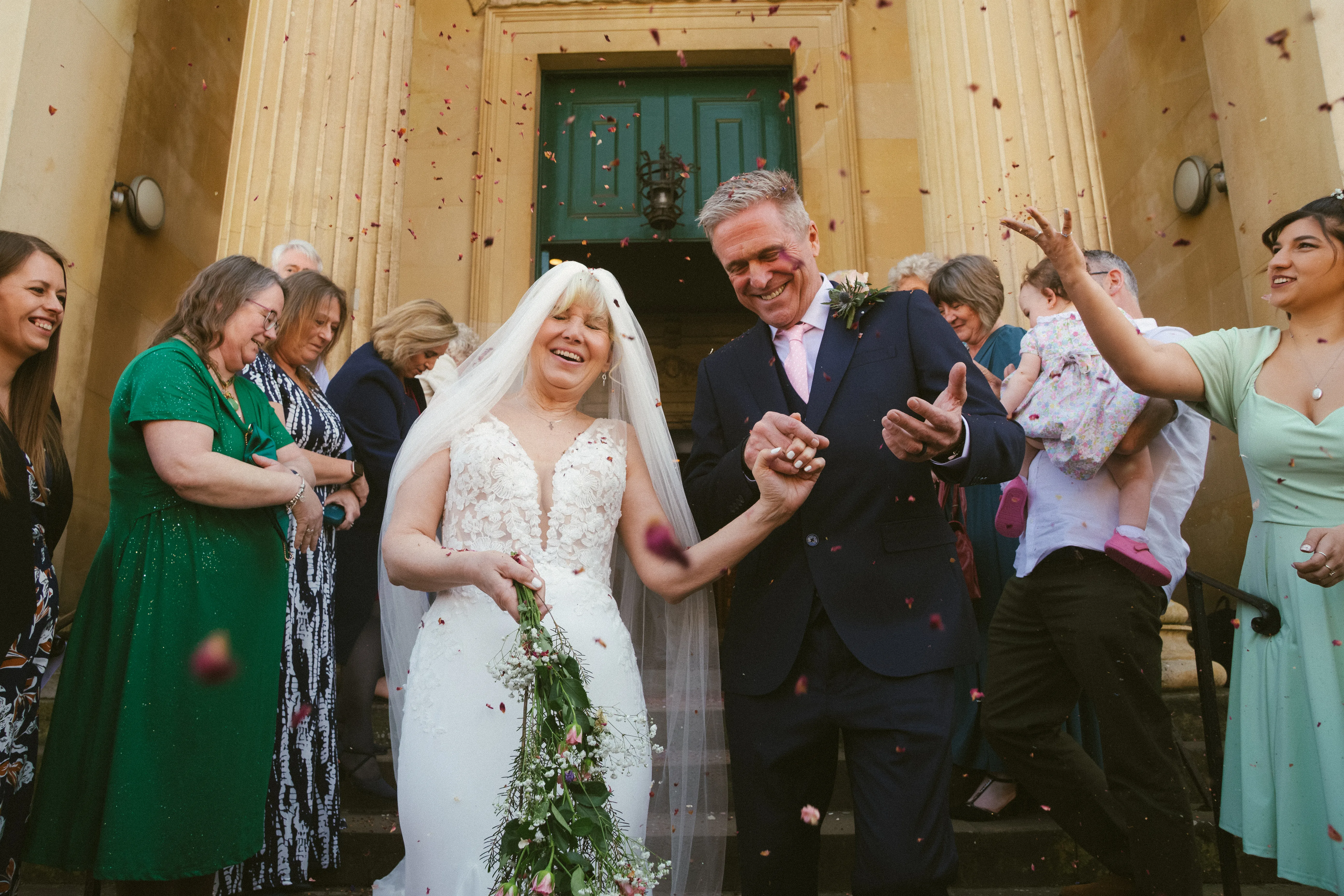 A newly married couple walking out through a shower of confetti, a green doorway behind them.