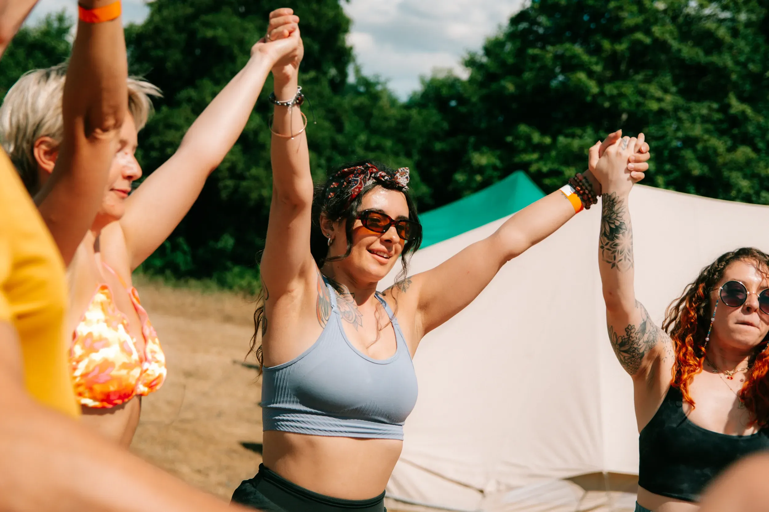 Women dancing in a circle with their hands held high, sunglasses on and a stretch tent behind them.