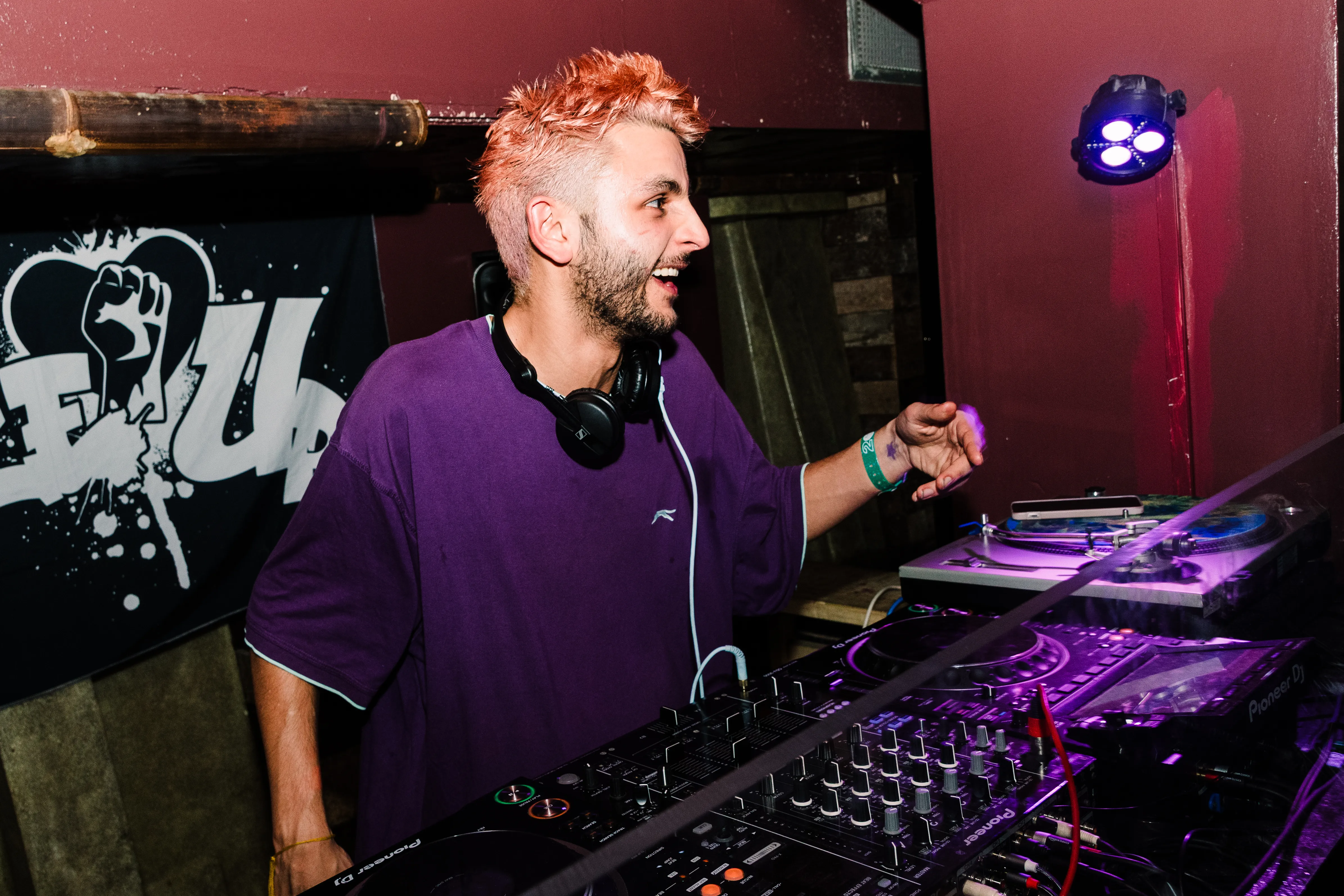 A DJ with orange hair in a purple shirt grinning behind the decks under pink light.