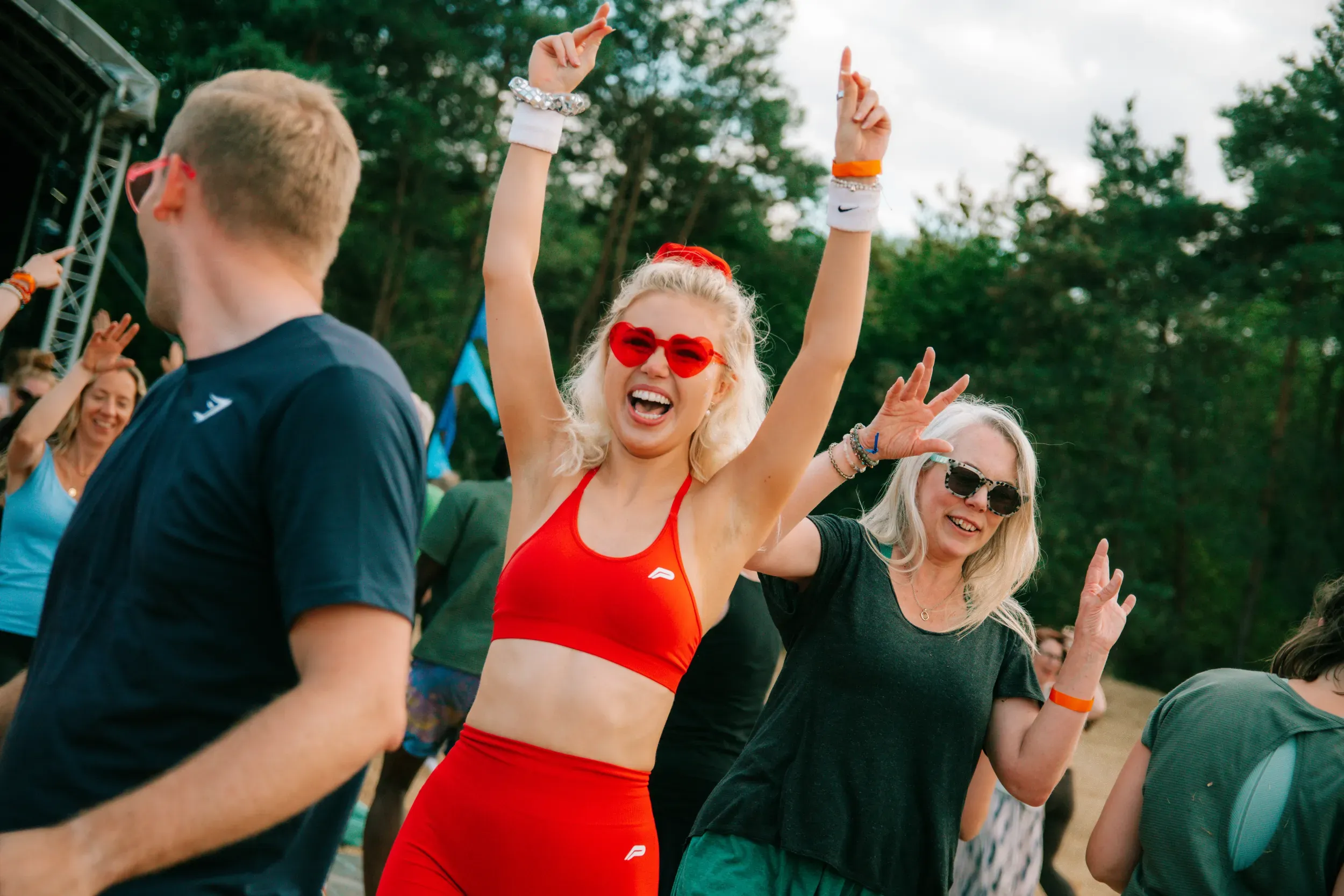 A woman in red heart-shaped sunglasses dancing with both hands in the air near the stage.