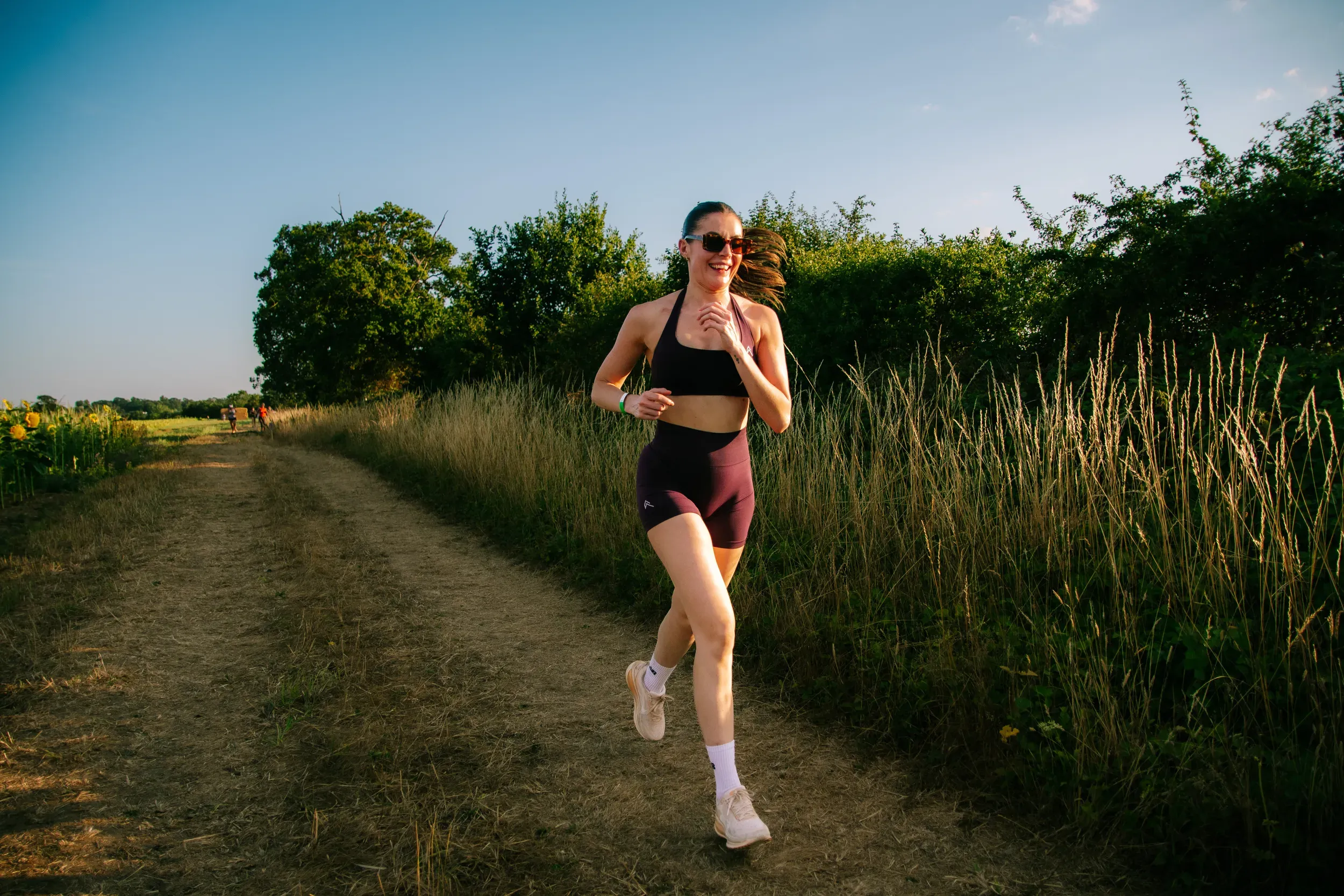 A runner in maroon coming along a dry track beside a tall hedgerow, low sun on her face.