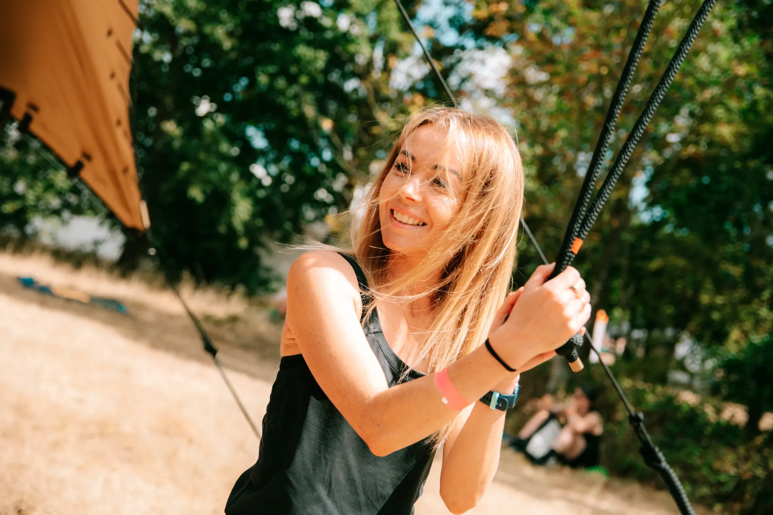 A woman laughing as she holds onto an aerial rope in the sunshine, canopy overhead.