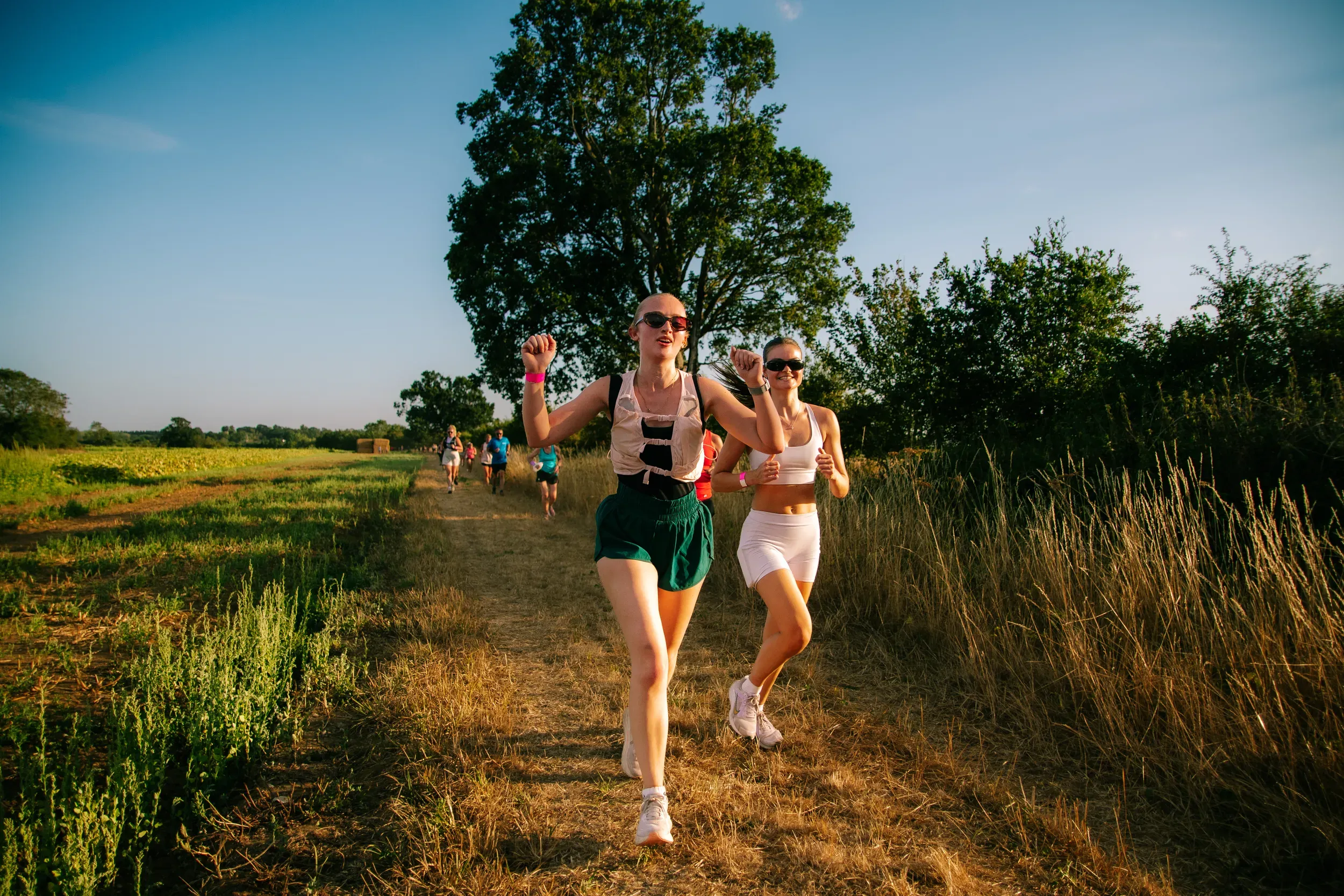 Two runners side by side on a dirt track under a big tree, more of the group spread out behind.