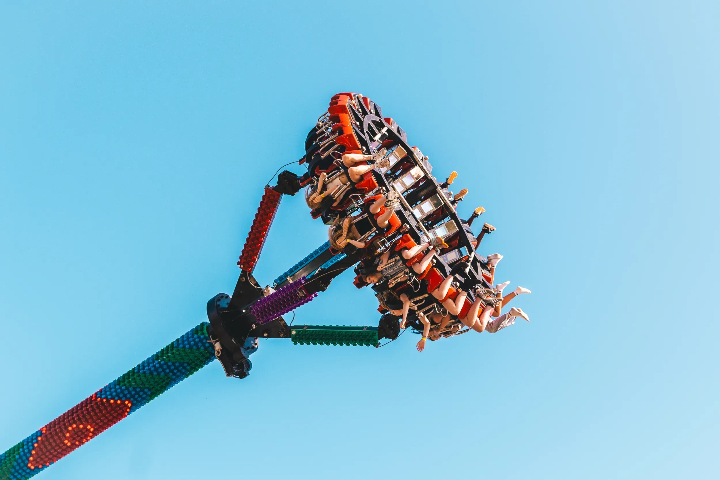 A fairground swing ride raised against a bright blue sky, riders strapped into its spinning arm.