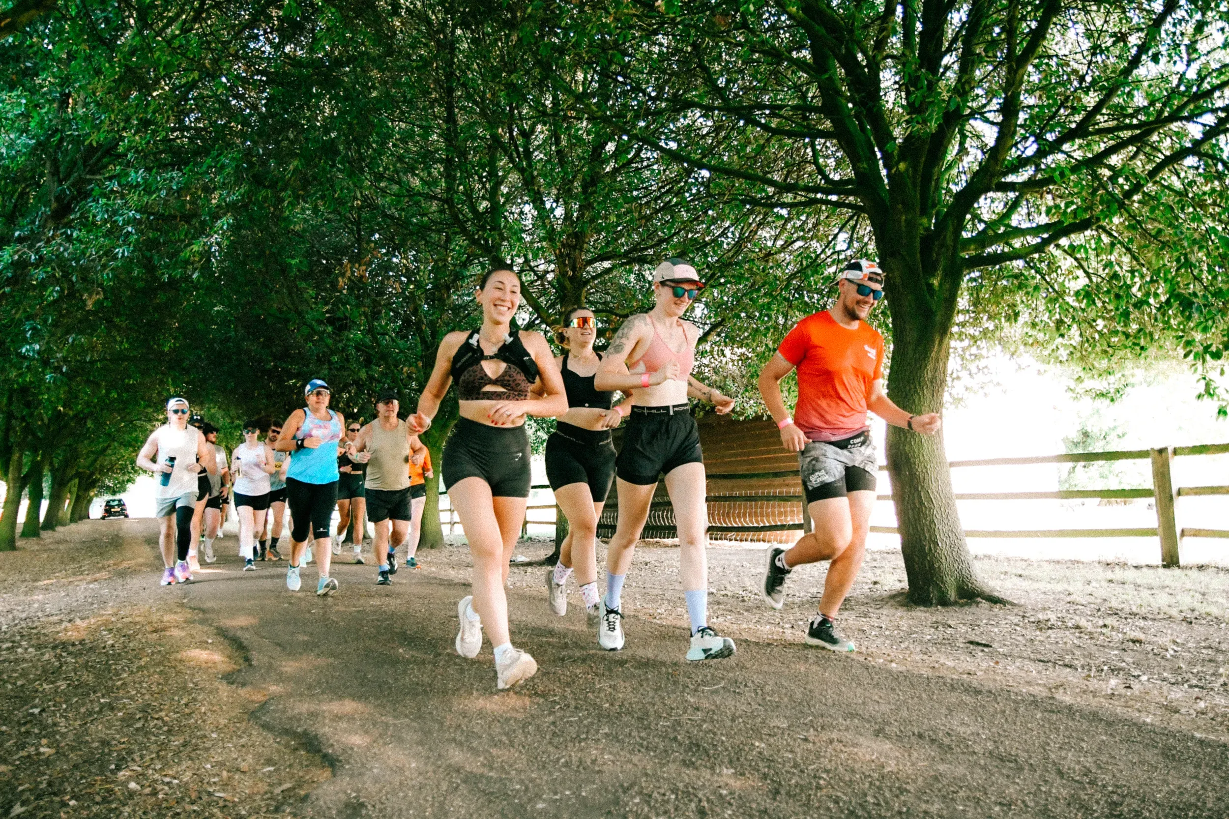 A pack of runners coming down a tree-lined track in dappled sunlight, front three grinning and holding hands.