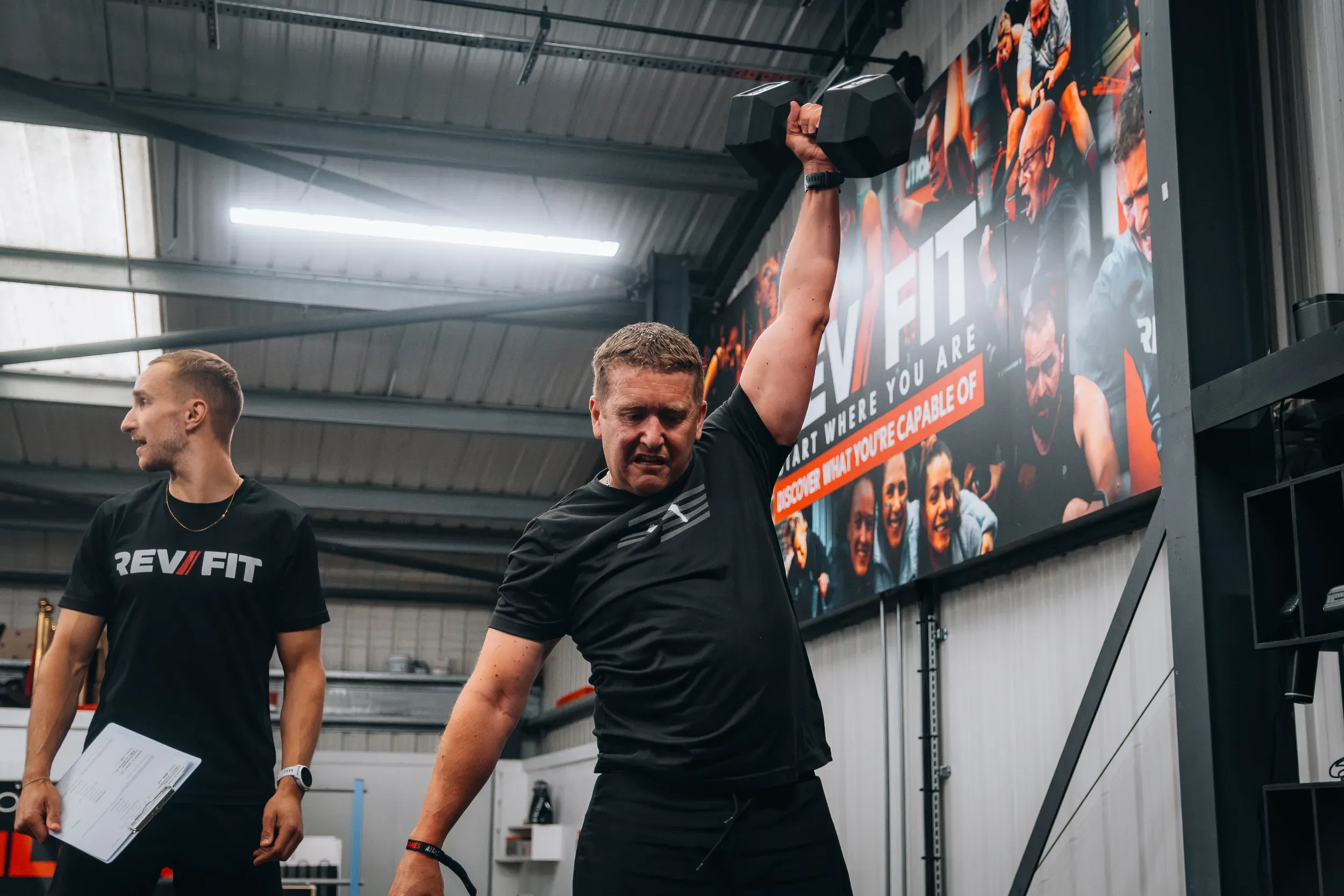 A judge with a scoresheet walking beside a competitor carrying a dumbbell at a gym event.