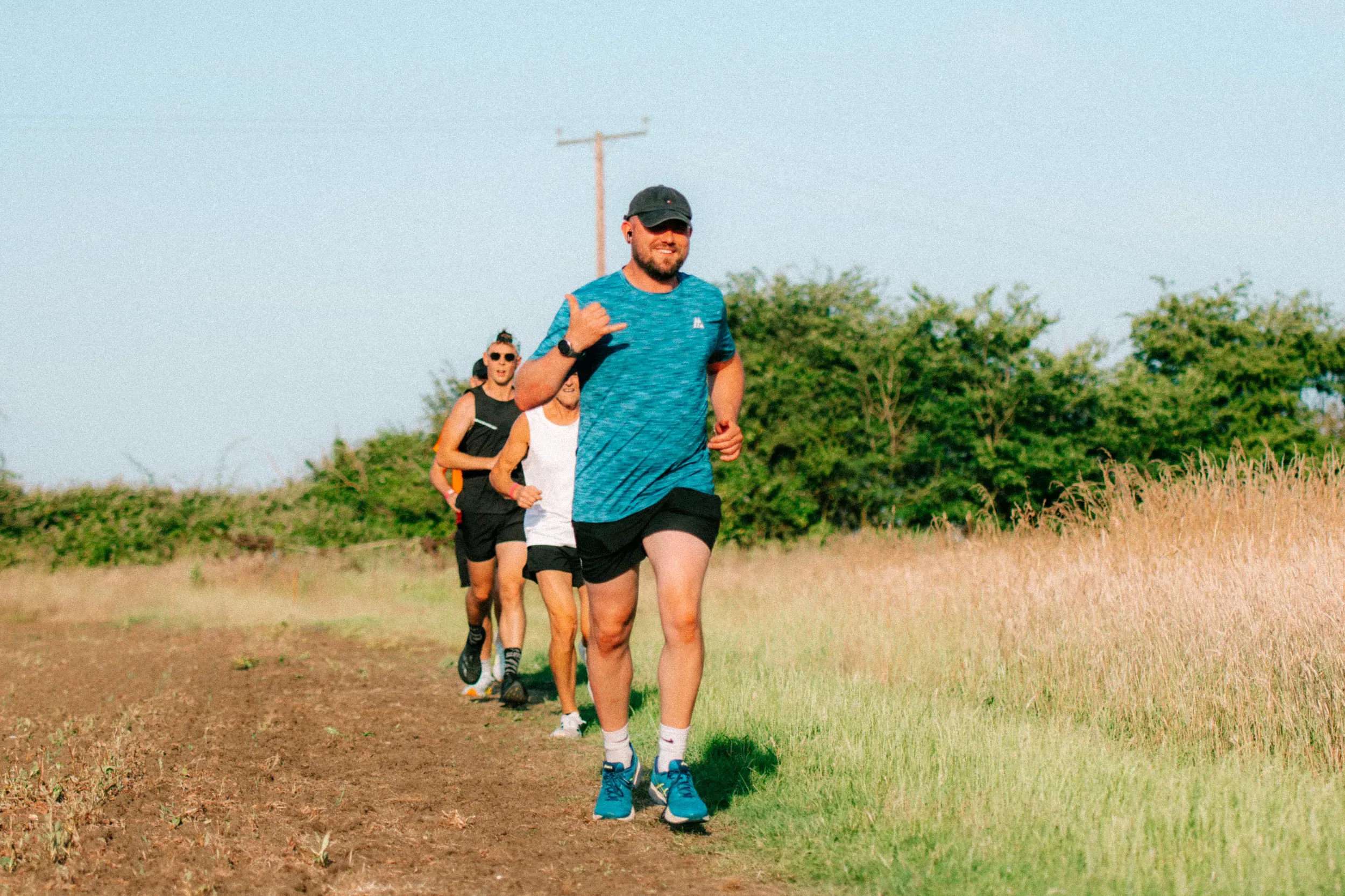 A runner in a teal top throwing a shaka at the camera, three more strung out behind him on the path.