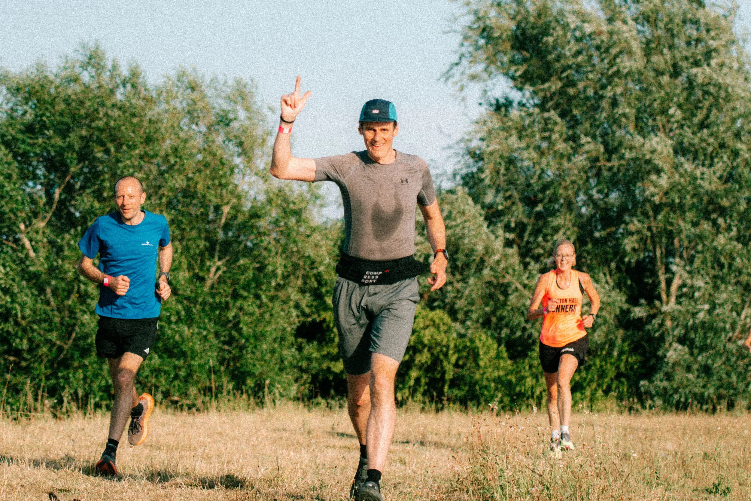 A runner holding up a peace sign mid-stride on a grass track, two others either side of him.