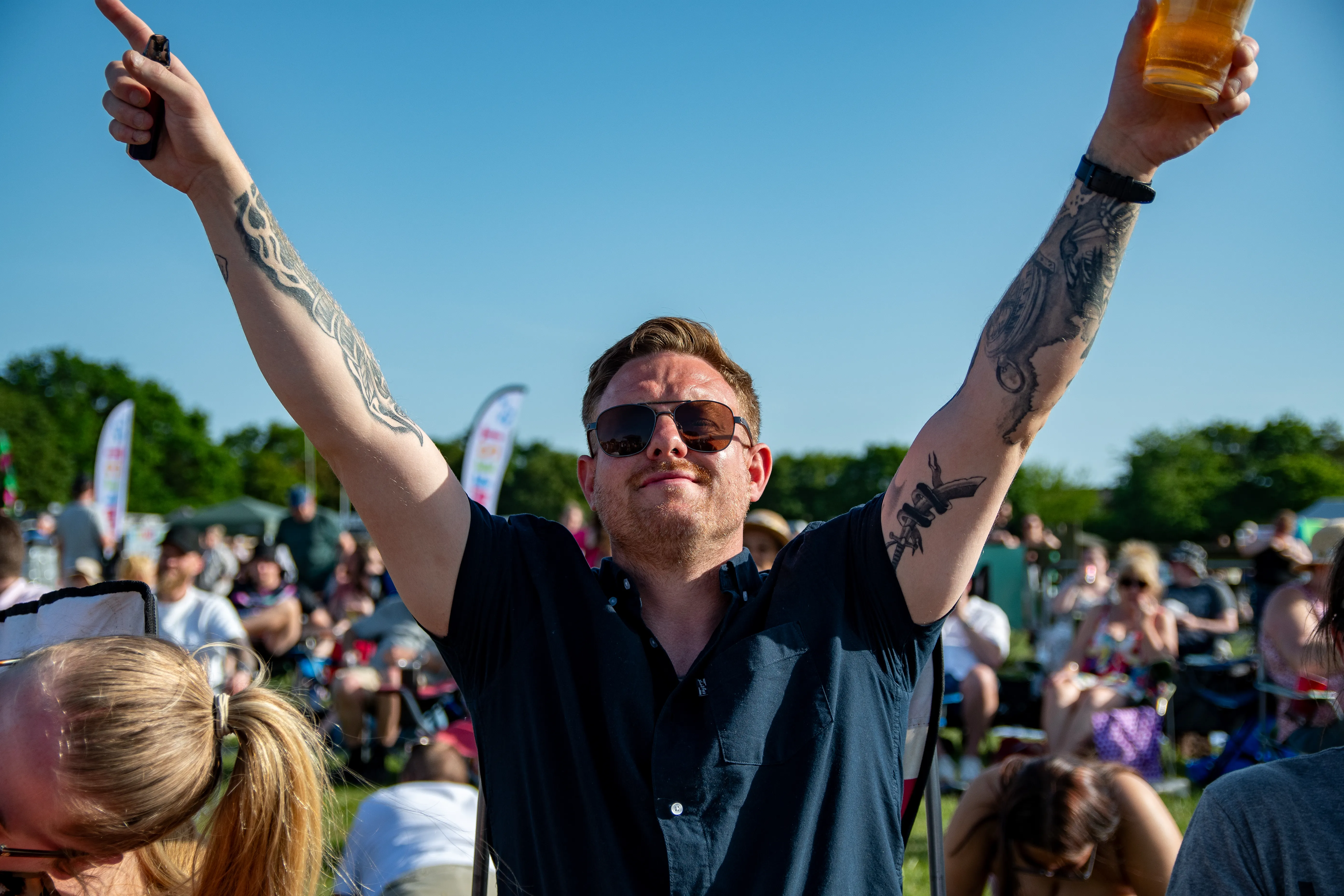 A man with tattooed arms aloft, drink in hand, in the crowd.