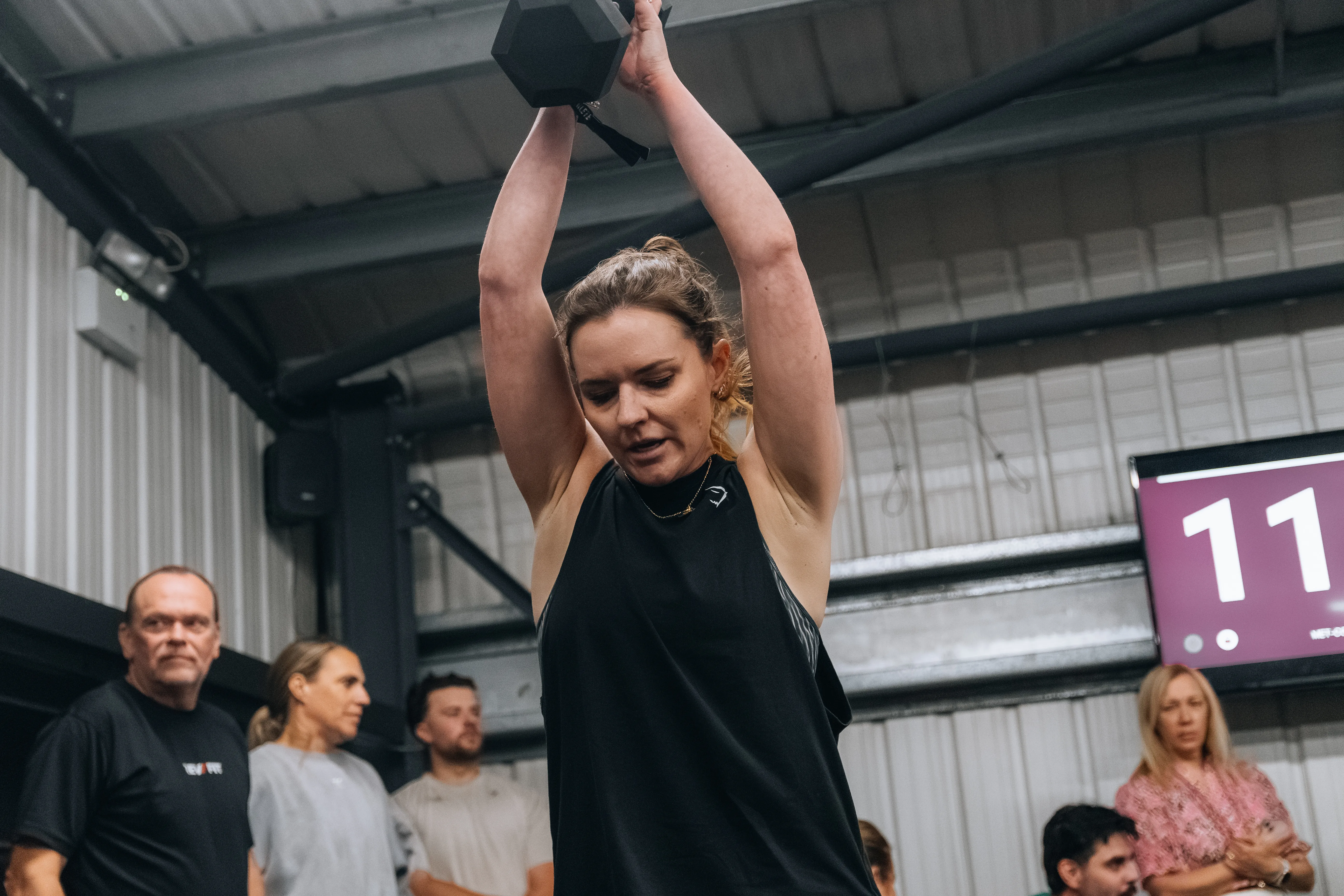 A competitor holding a dumbbell locked out overhead beside the scoreboard.