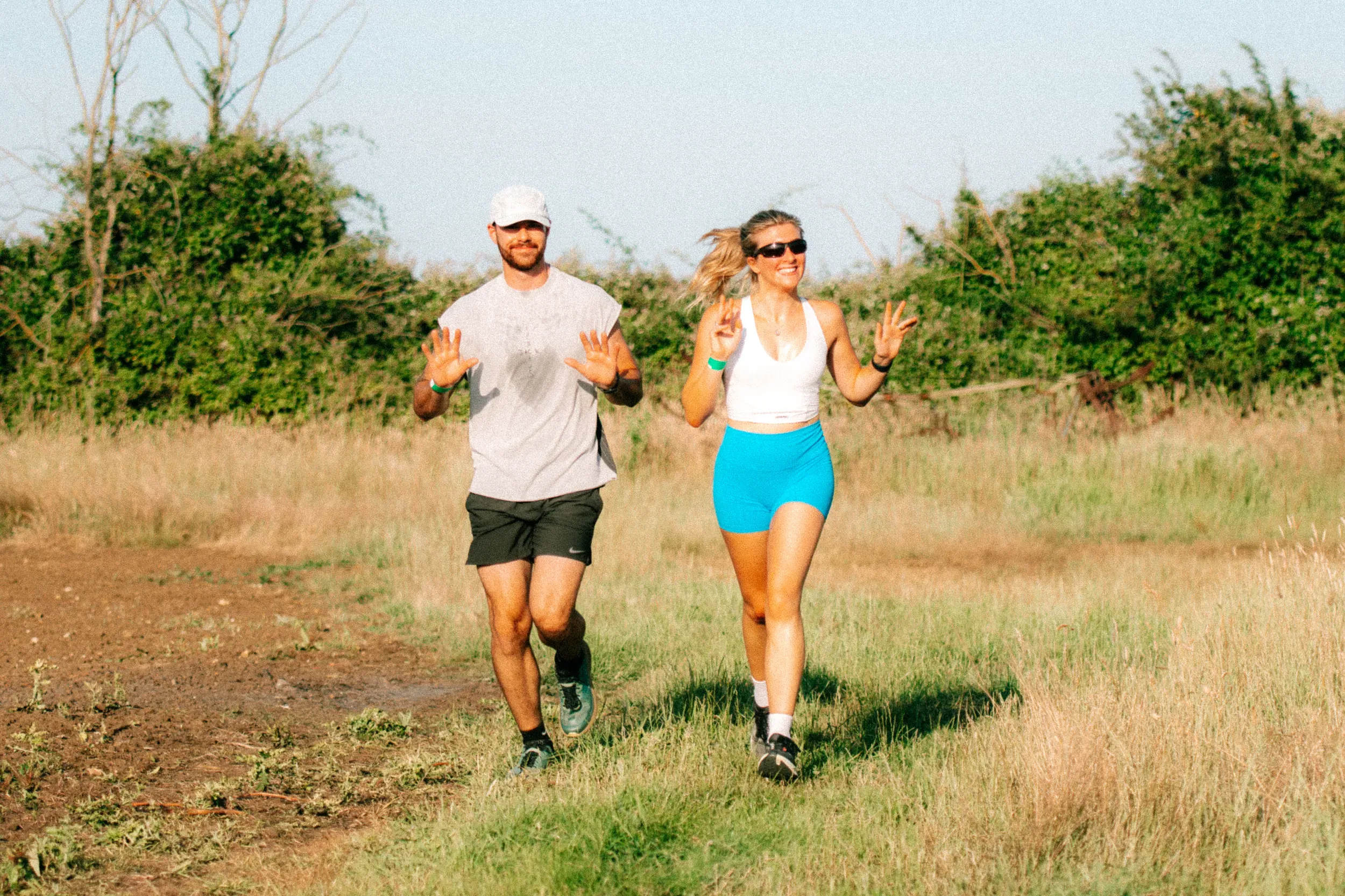 A man and a woman running side by side with both hands raised in a wave, hedgerow behind them.