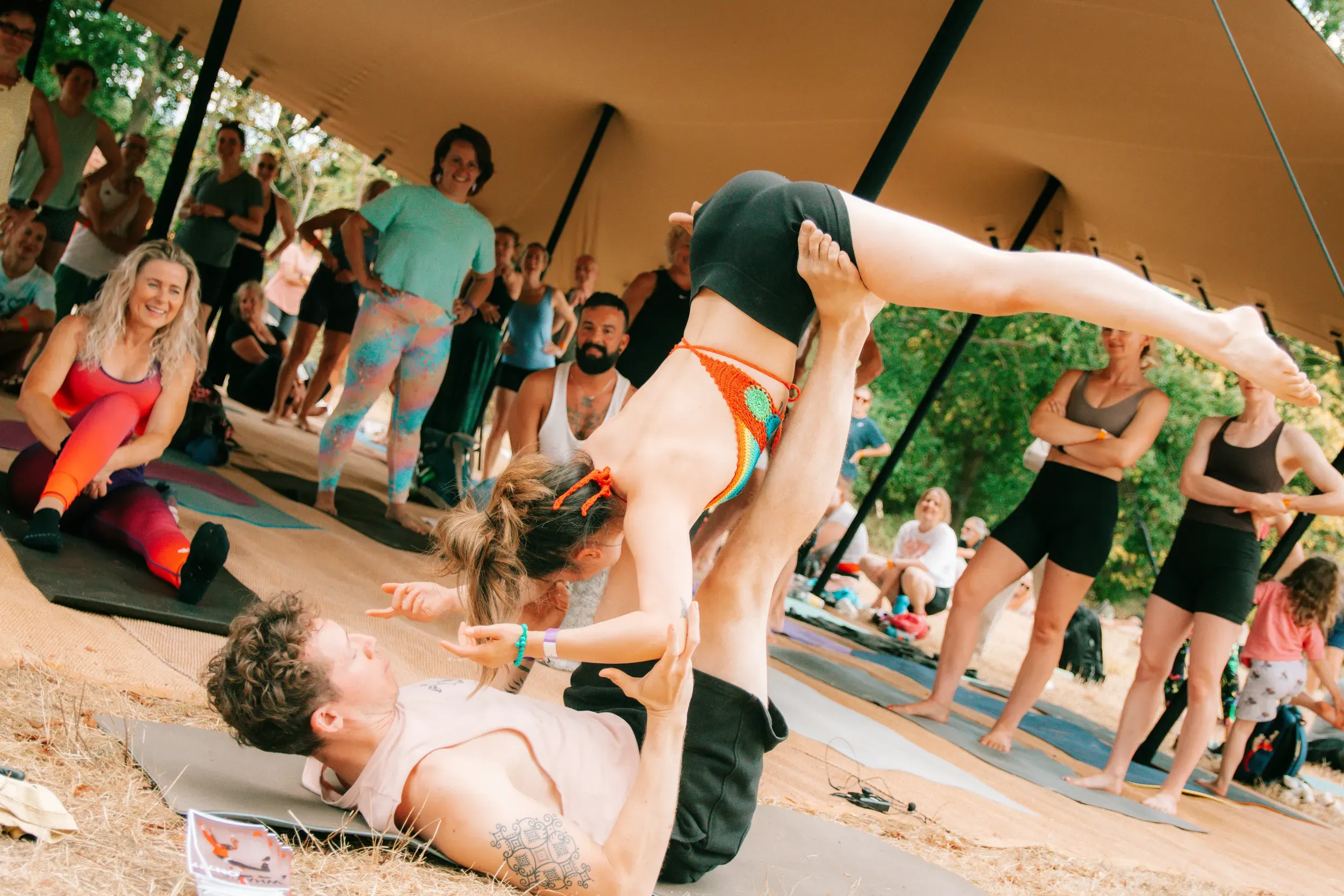 An acroyoga pair mid-pose on the mat while the rest of the class watches from around the tent.