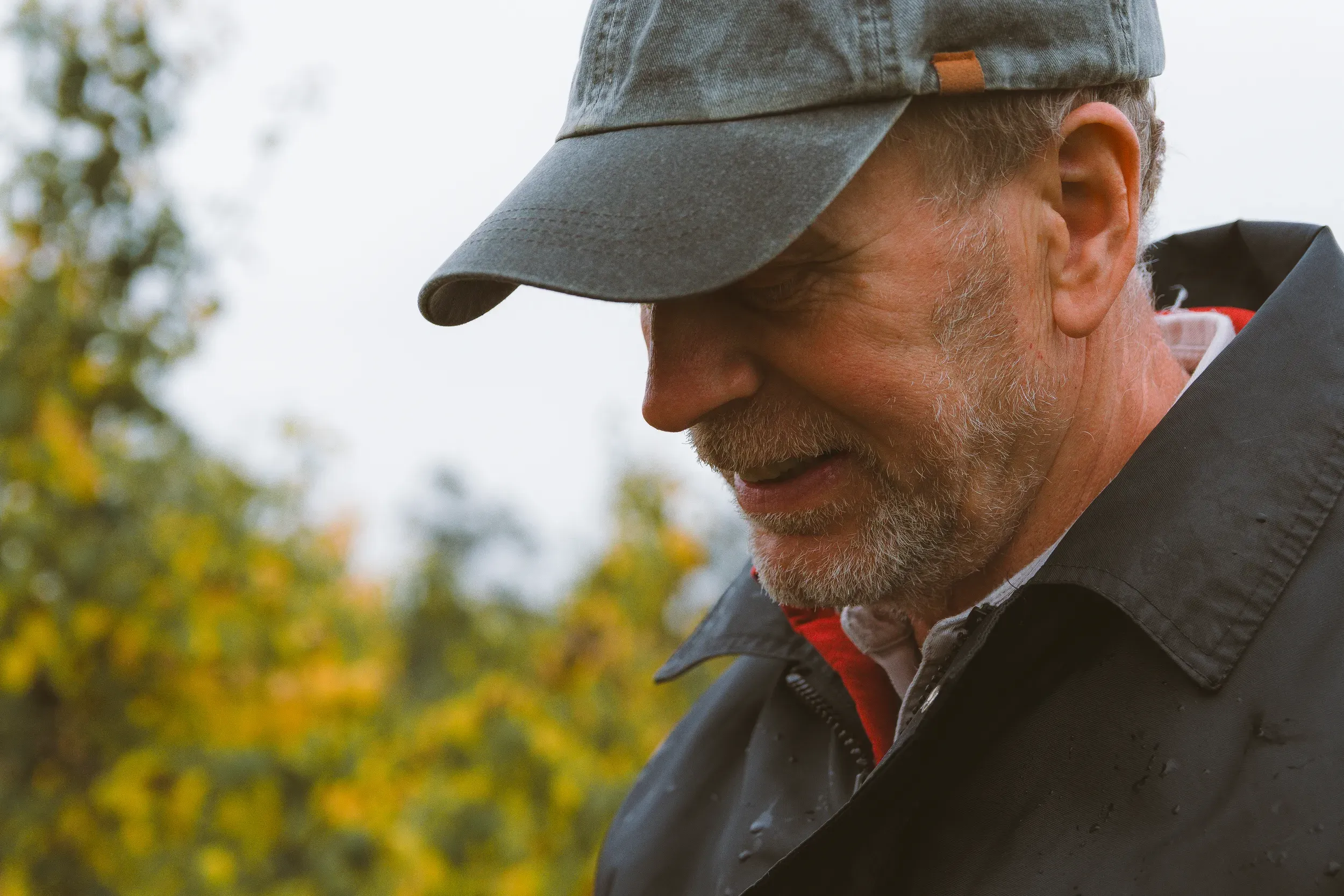 A close profile of an older man in a flat cap, autumn foliage soft behind him.