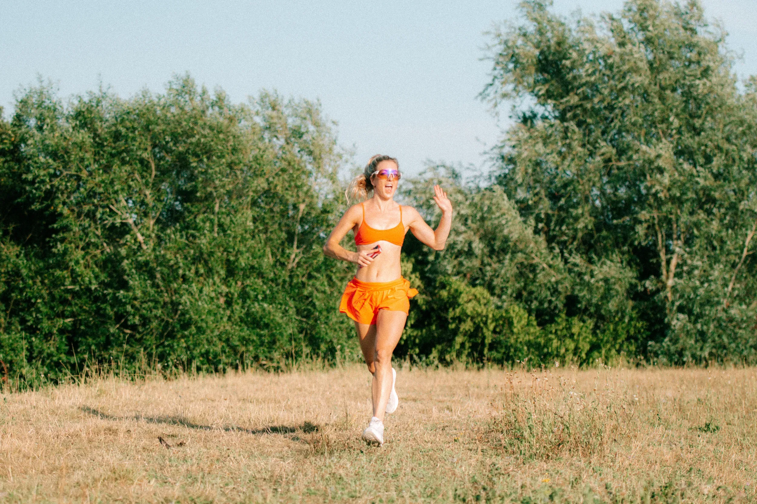 A runner in bright orange waving as she comes across the meadow in low evening sun.