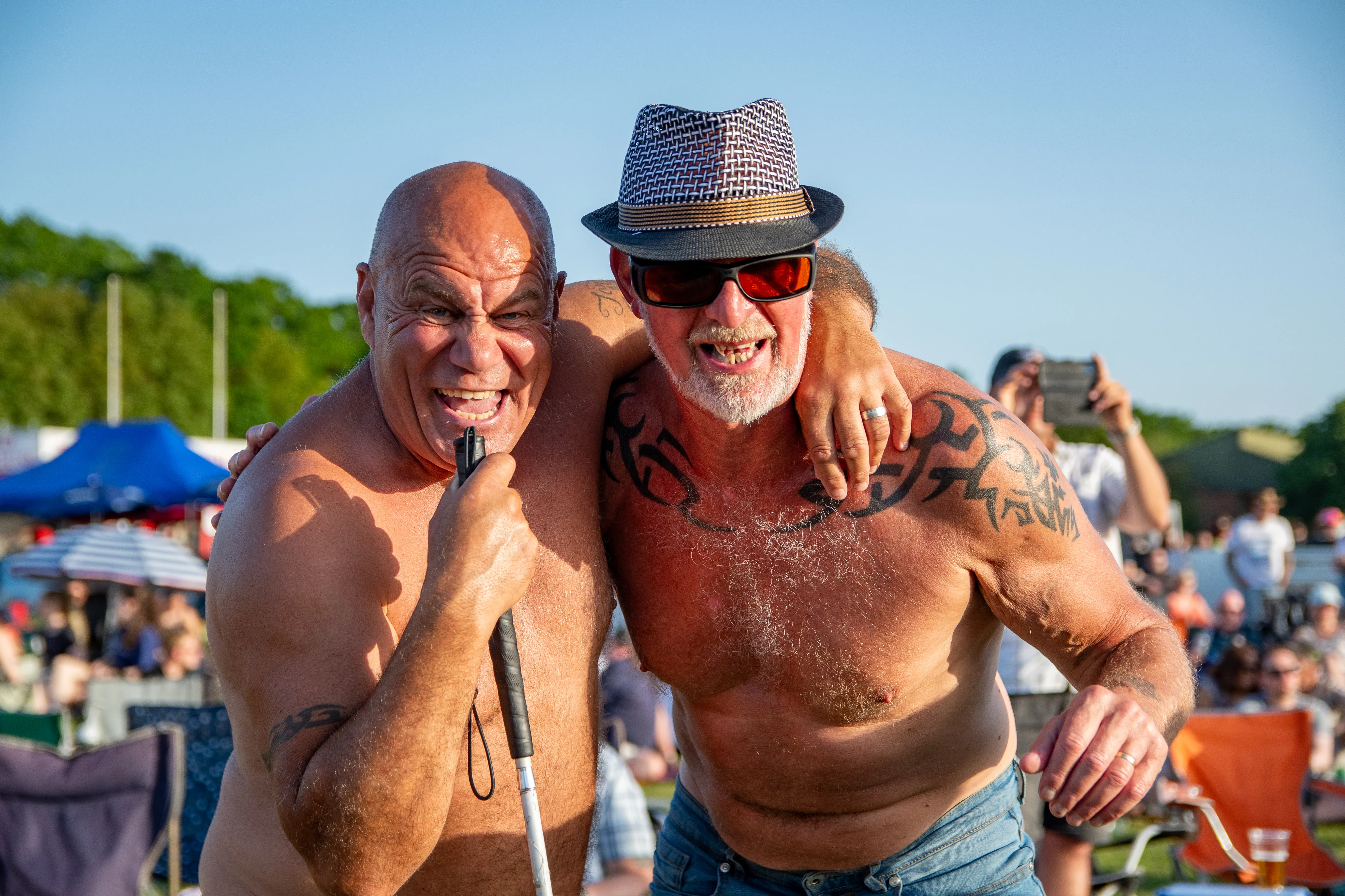 Two shirtless men in sun hats singing into a microphone together at a festival.