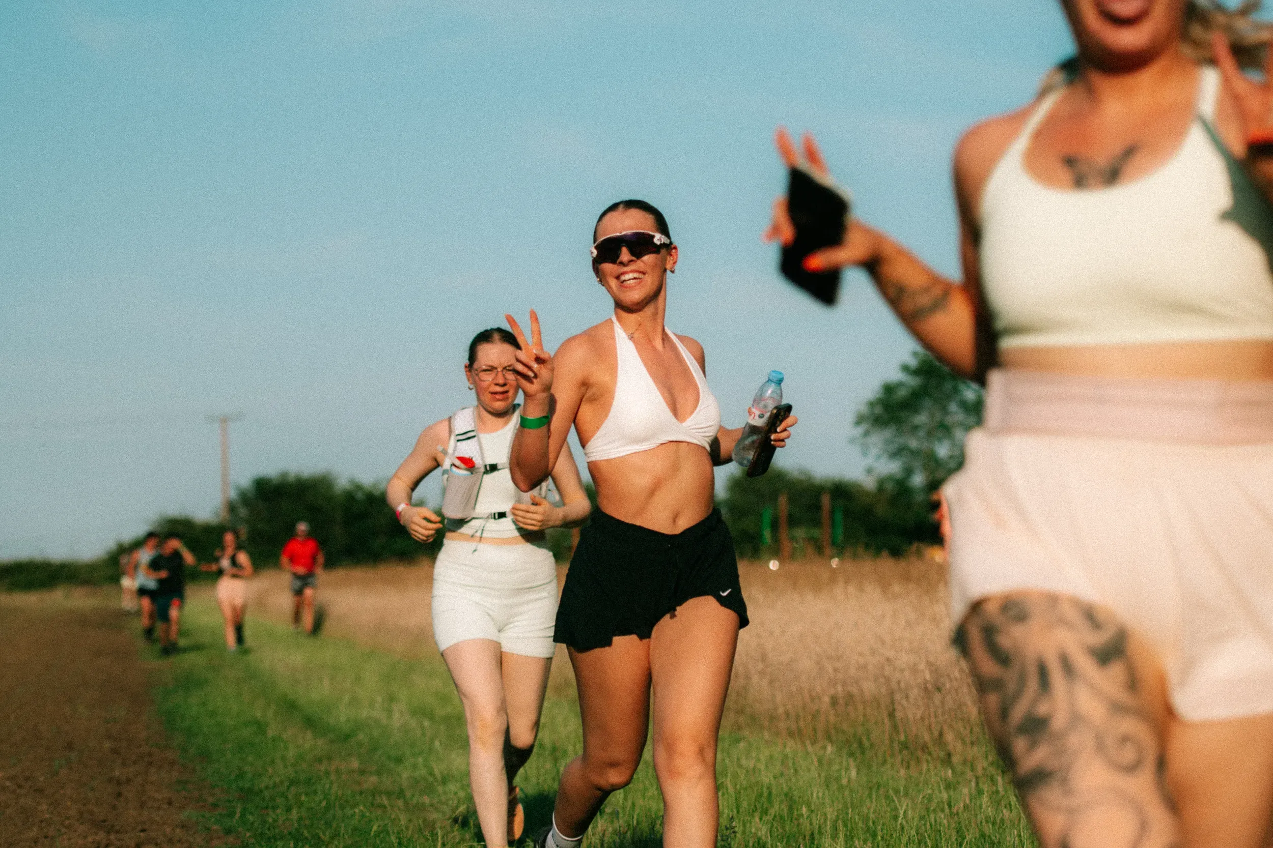 A runner in a white crop top flashing a peace sign, water bottle in hand, the group bunched up behind her.