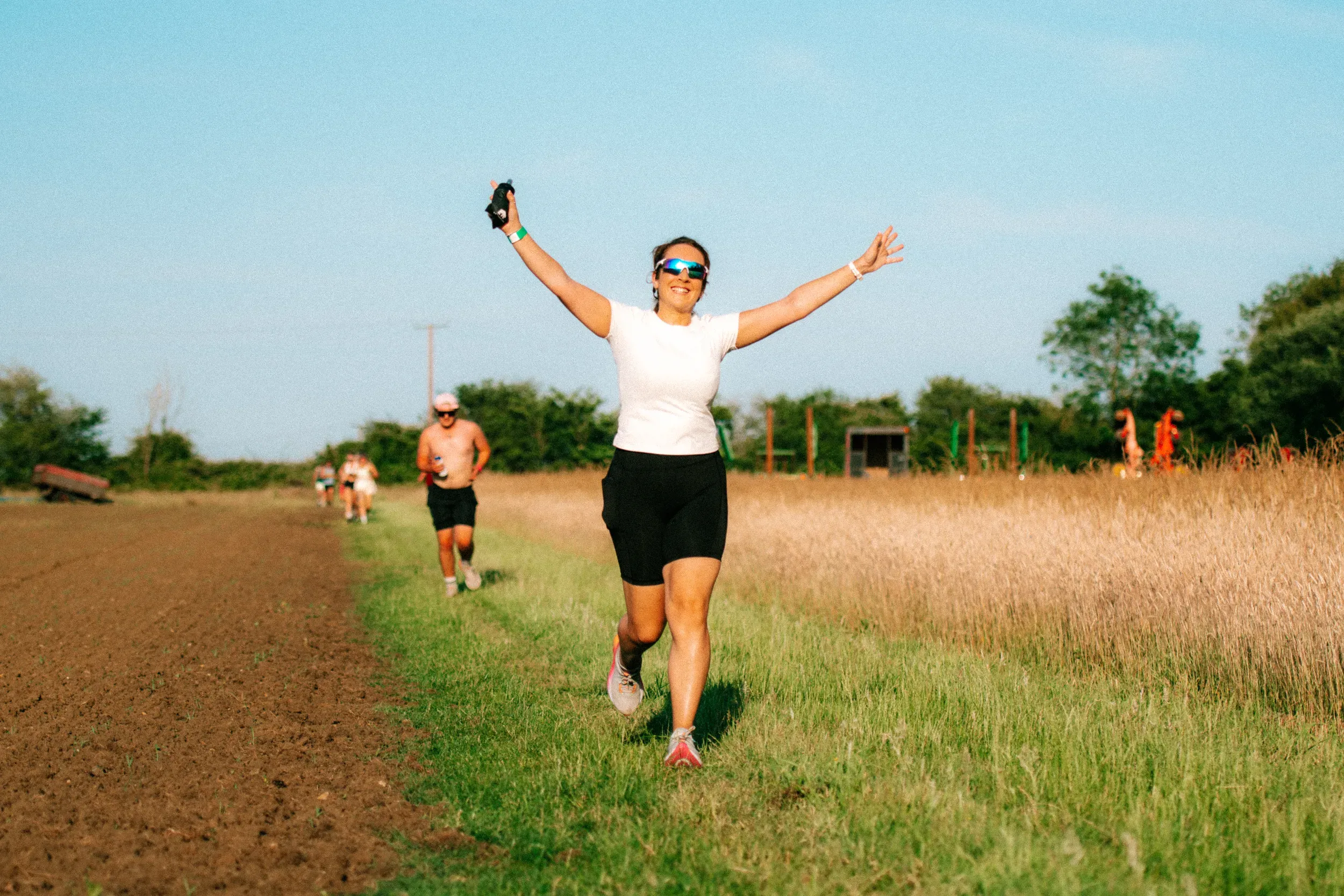 A runner throwing both arms wide and grinning as she comes up the grass strip beside a ploughed field.