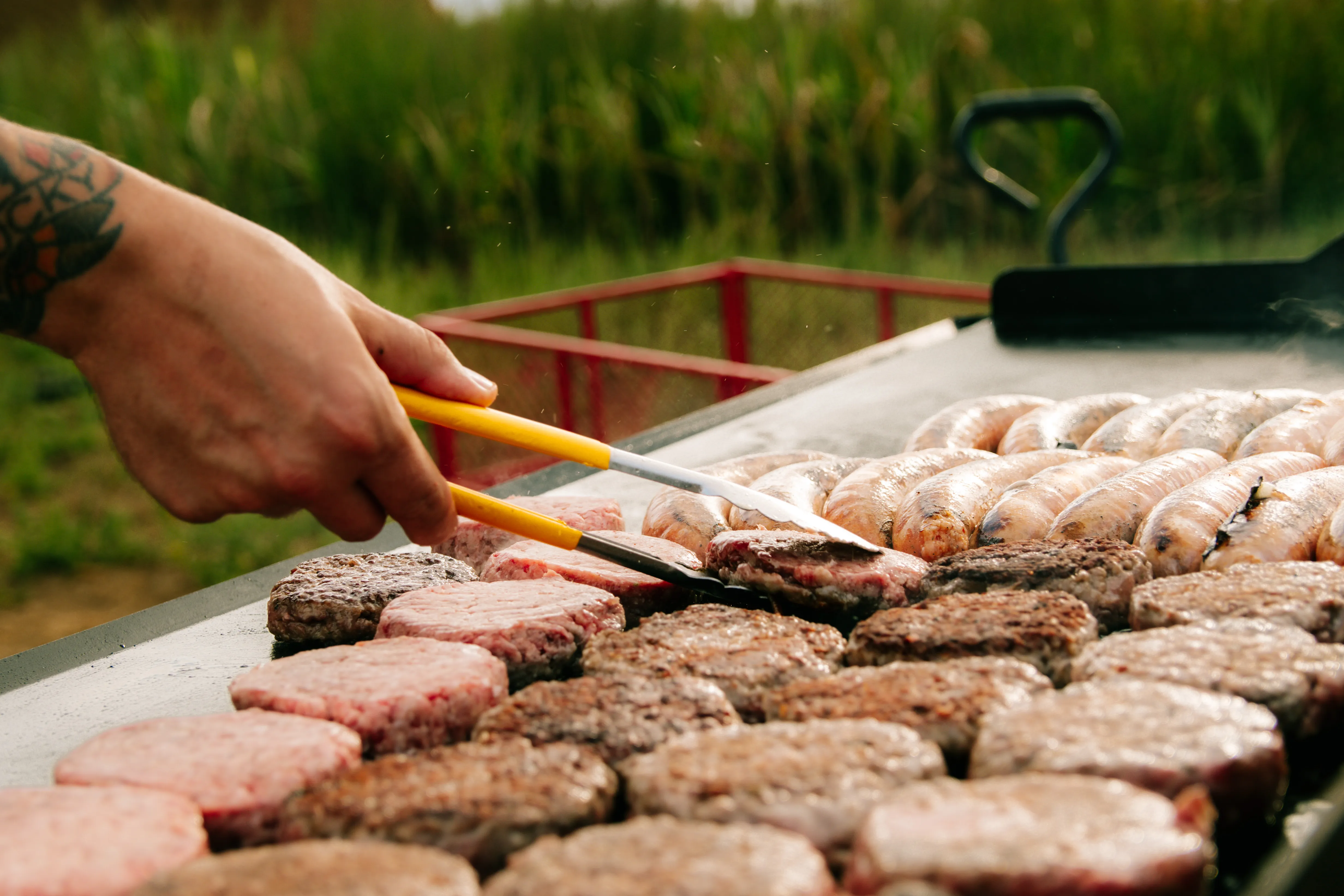 A tattooed arm turning burgers and sausages on the griddle beside the sunflower field.