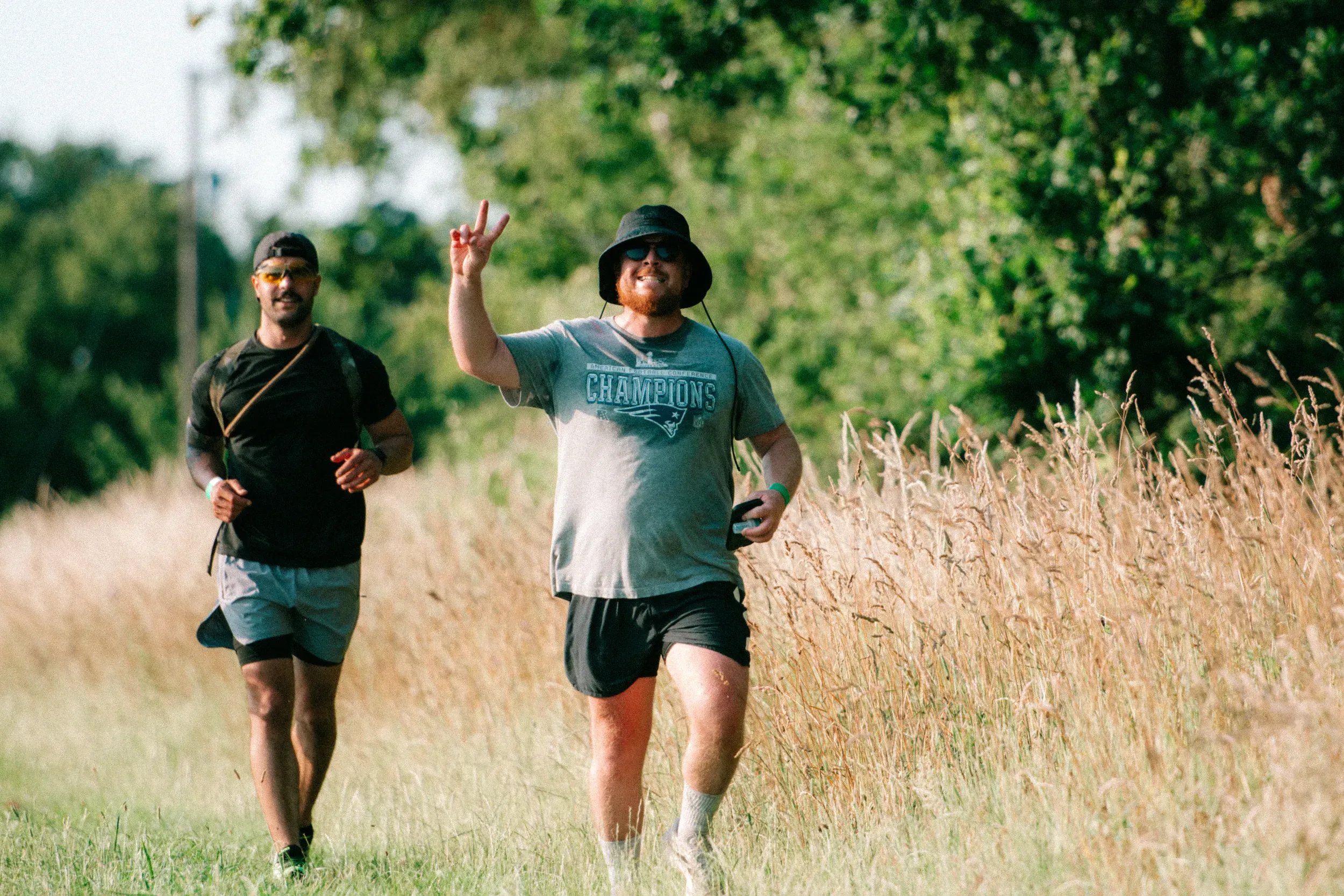 A runner in a bucket hat holding up a peace sign, his mate keeping pace beside him in the long grass.