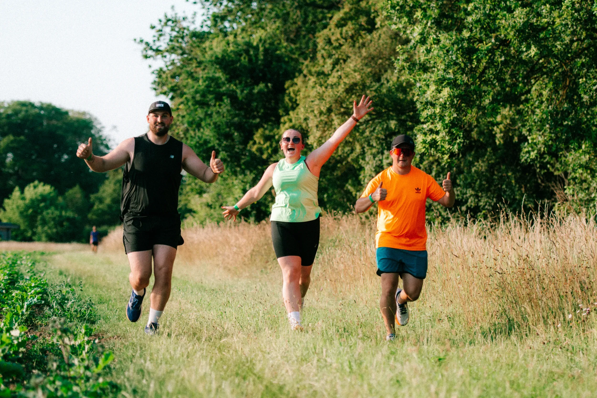 Three runners abreast on a grass path, thumbs up and arms out, all grinning at the camera.