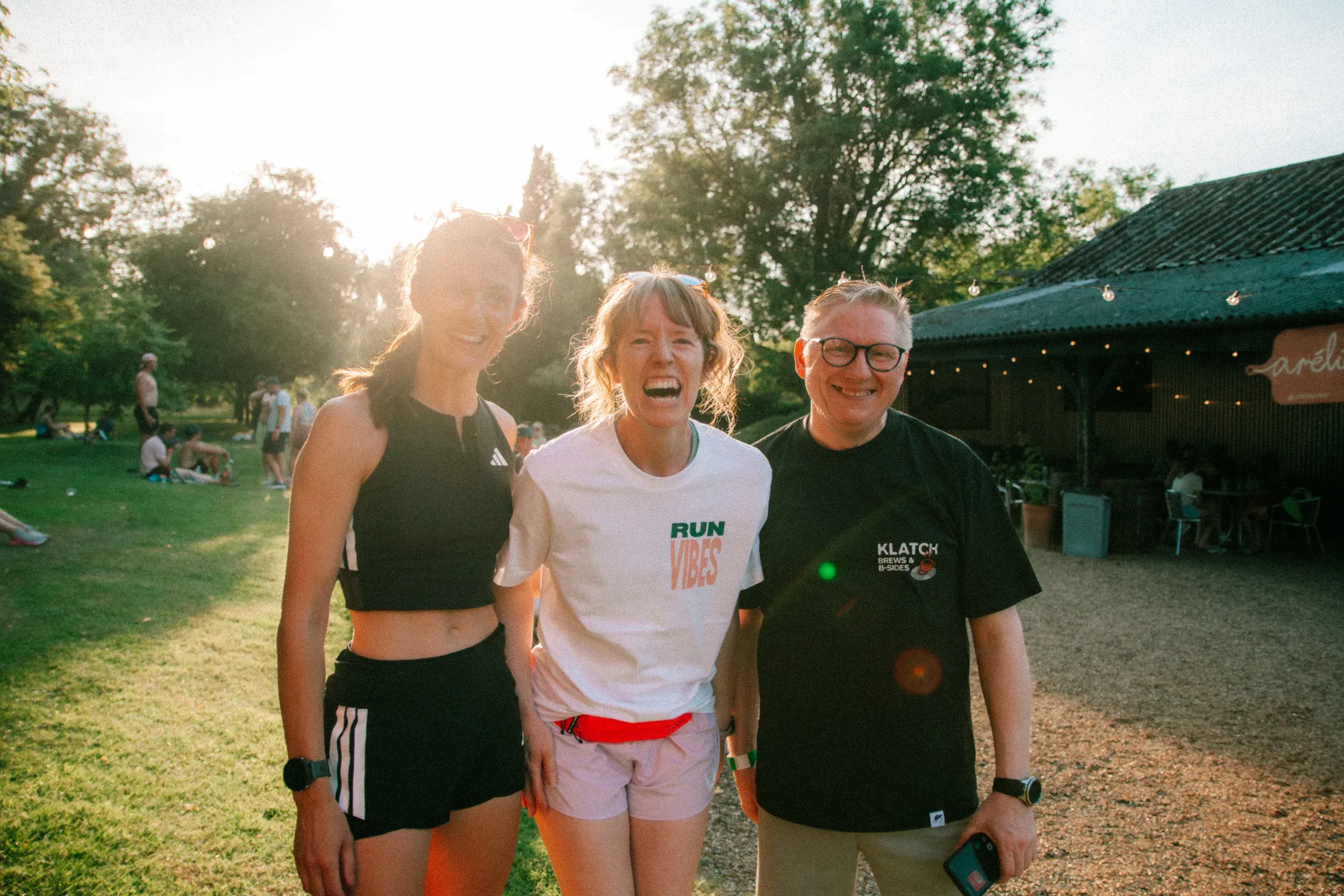 Three runners arm in arm outside the bar after the run, backlit by low sun, festoon lights strung along the barn behind them.
