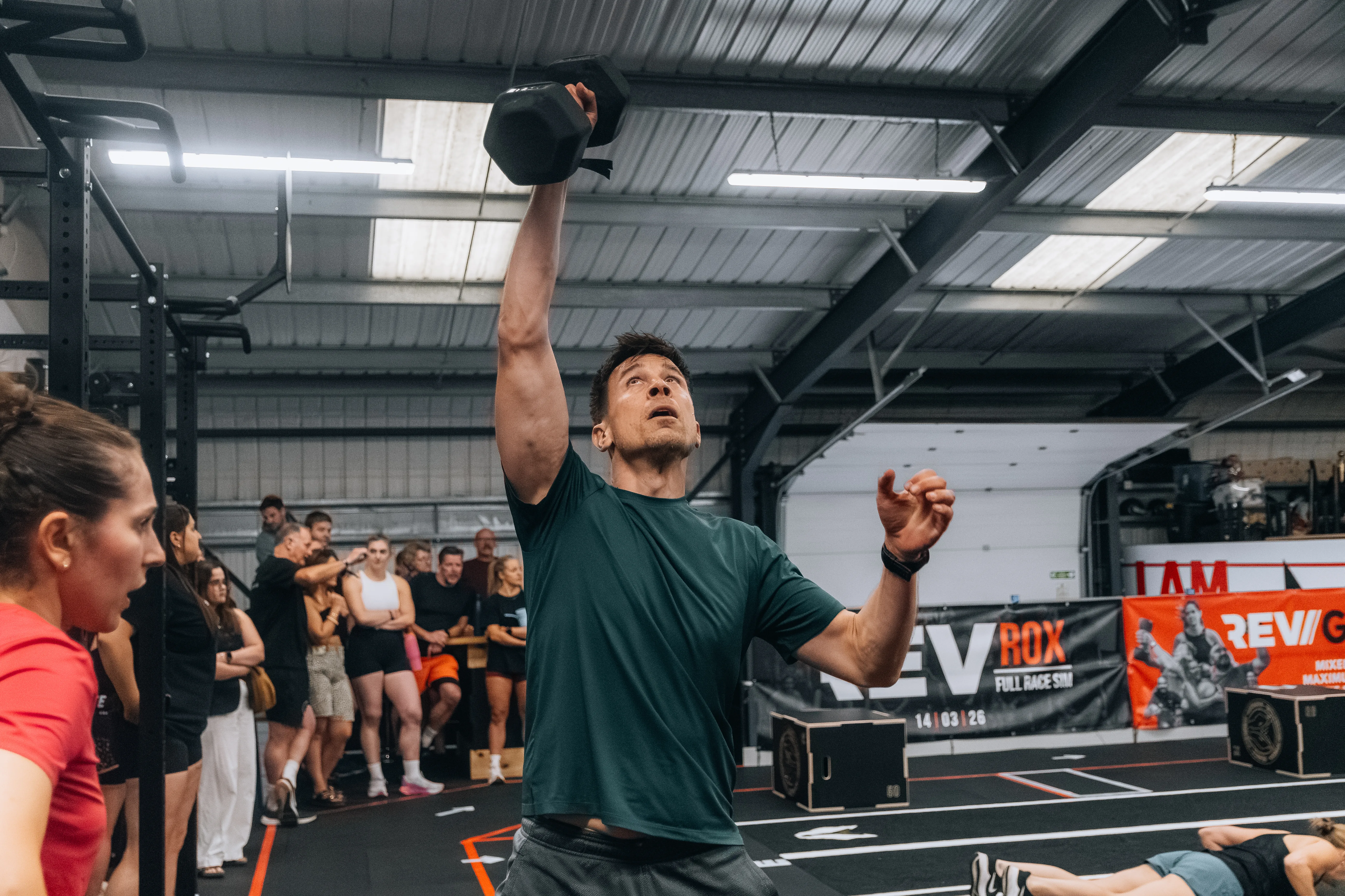 A competitor in green punching a dumbbell overhead mid-workout.