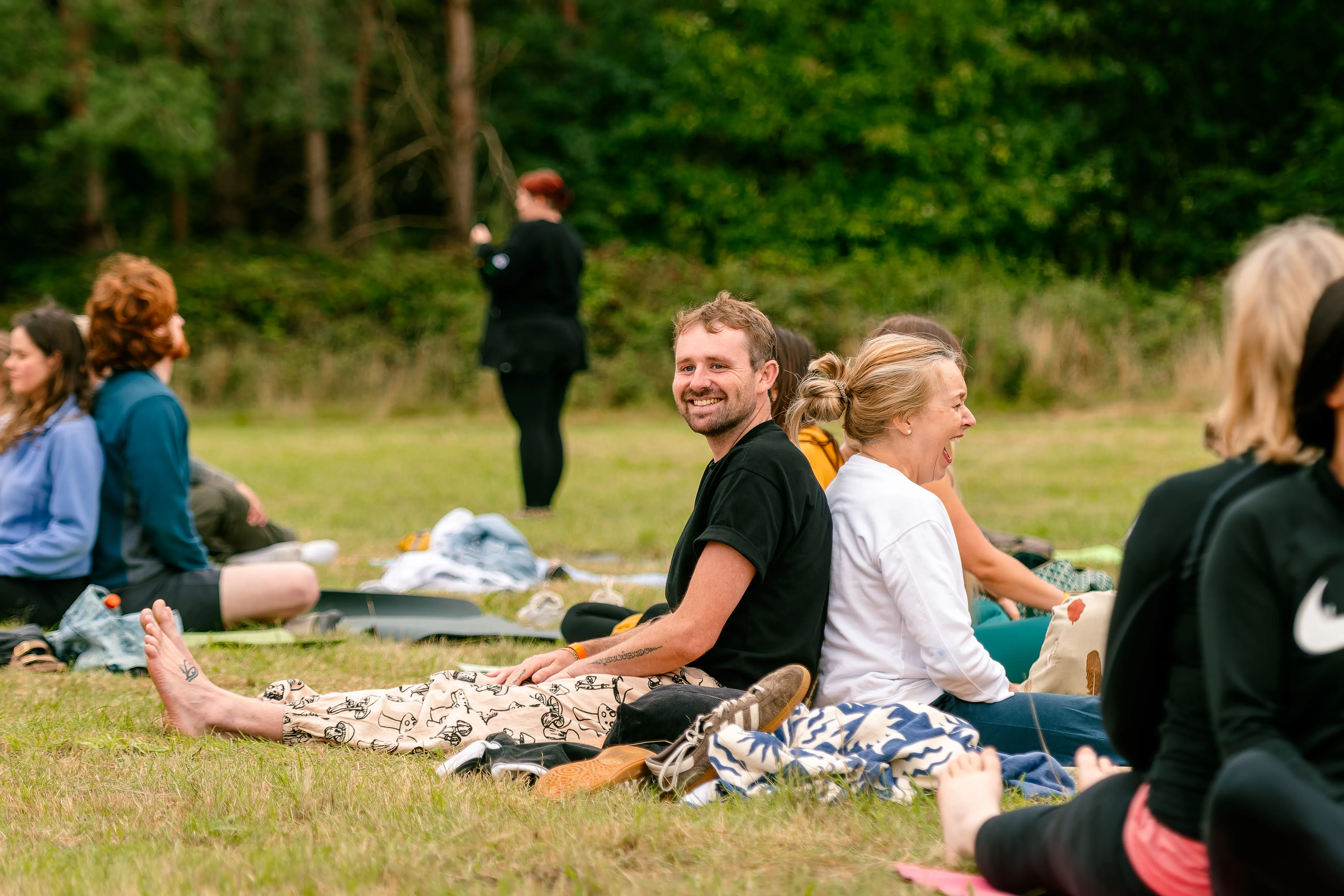A man and a woman sitting back to back on the grass, both laughing during a partner exercise.