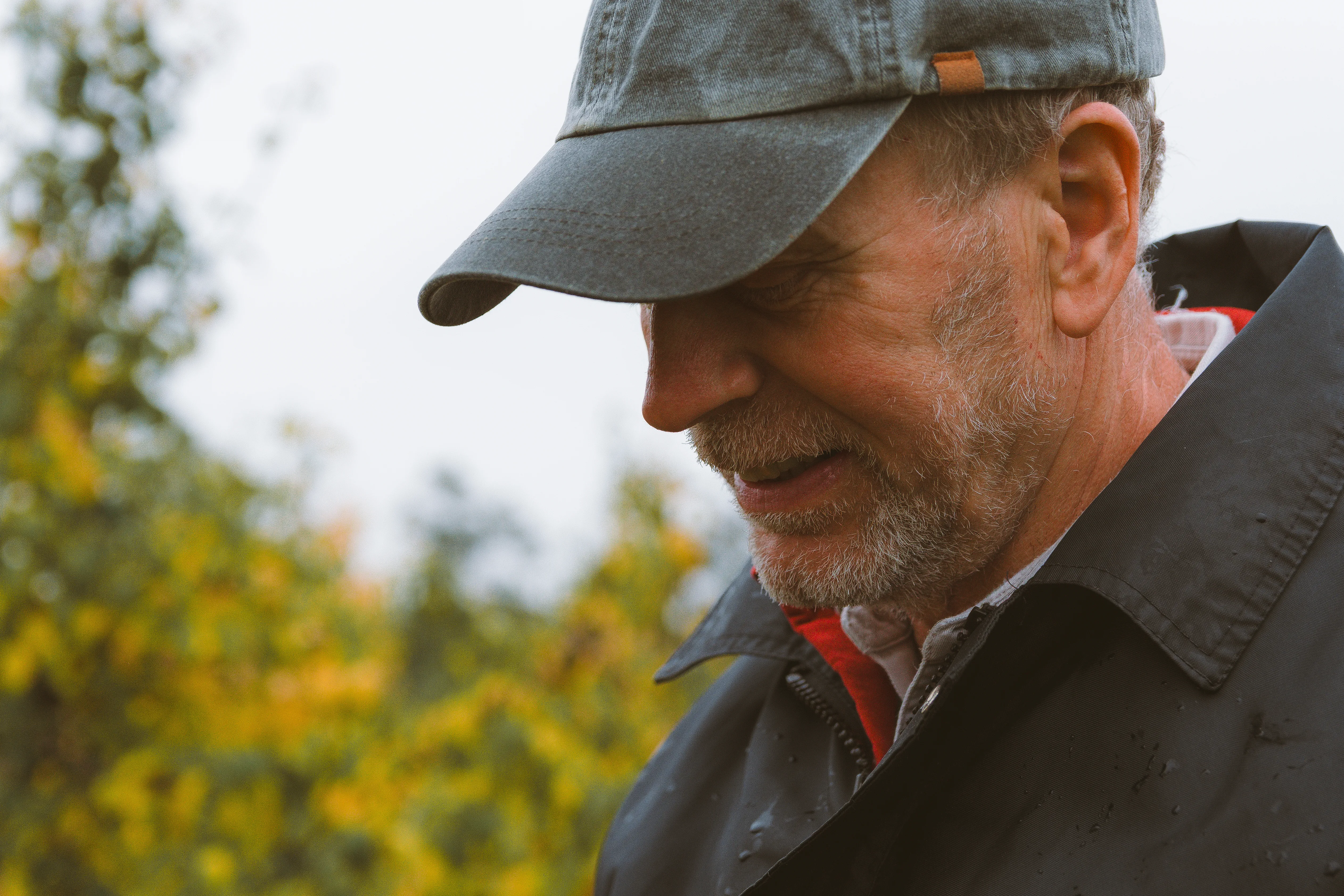 A close profile of an older man in a flat cap, autumn foliage soft behind him.
